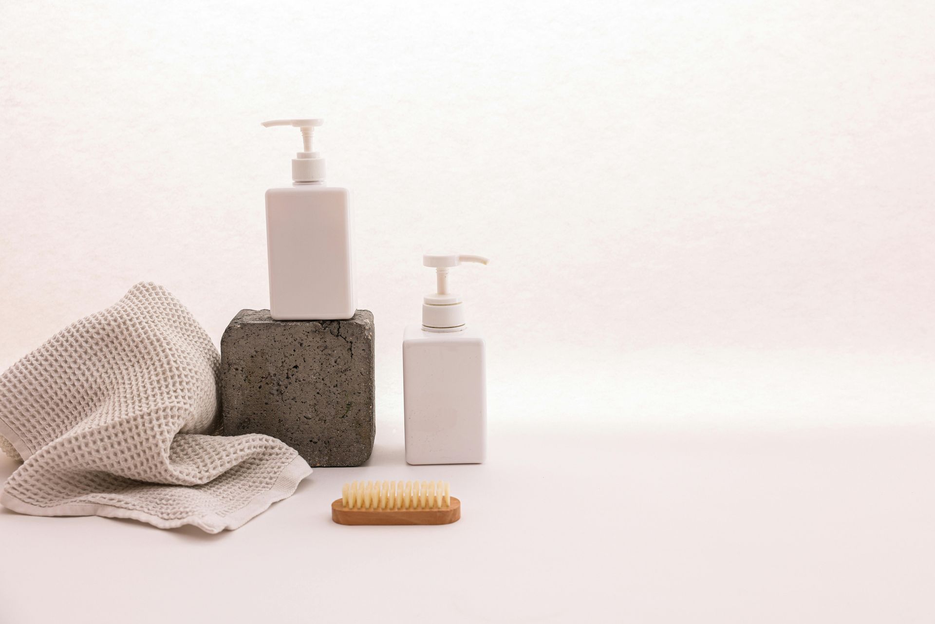 Two white soap dispensers, towel, and brush on a white surface, against a neutral backdrop.