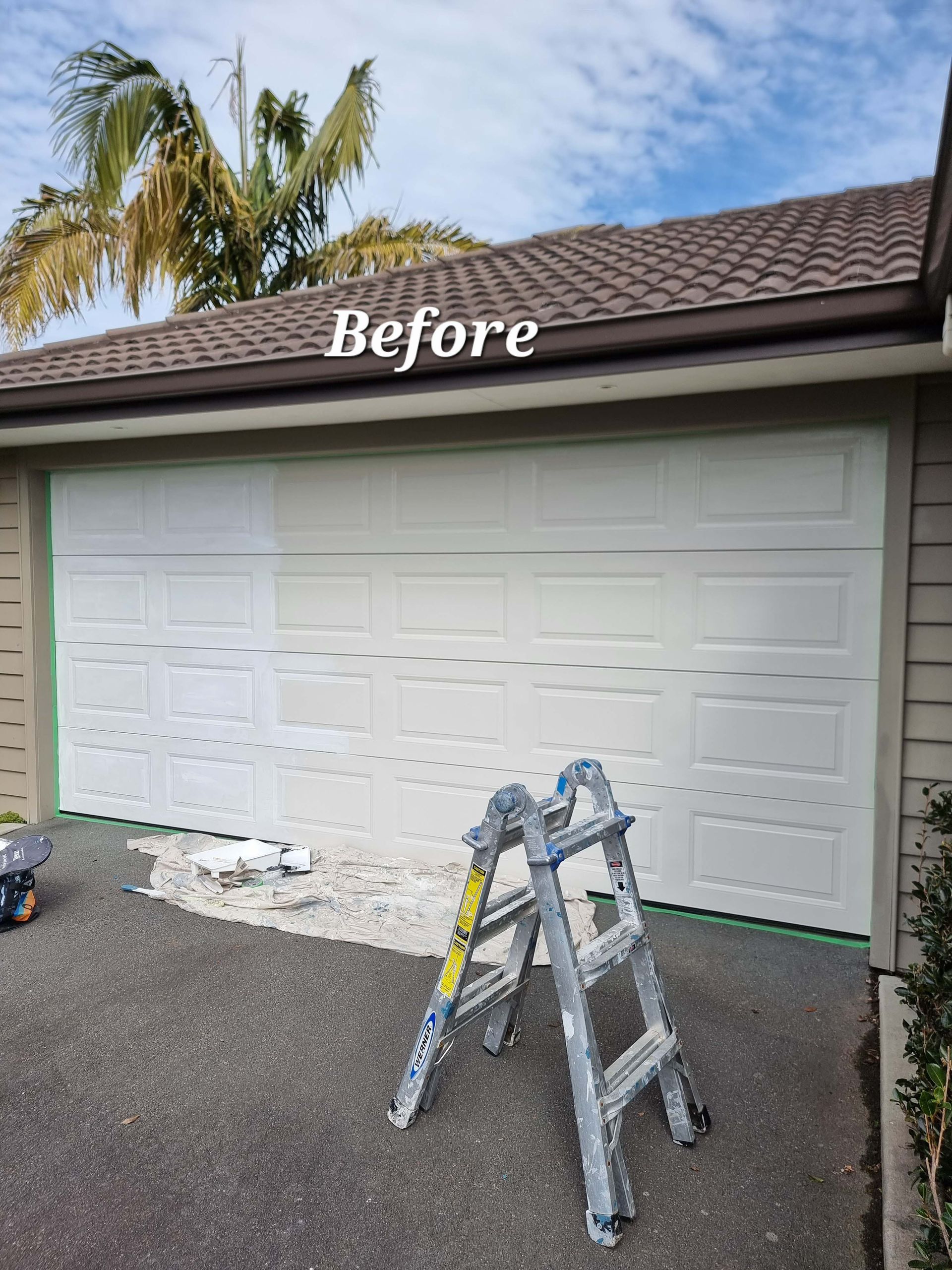 Before photo of a white garage door with a ladder in front and palm trees in the background