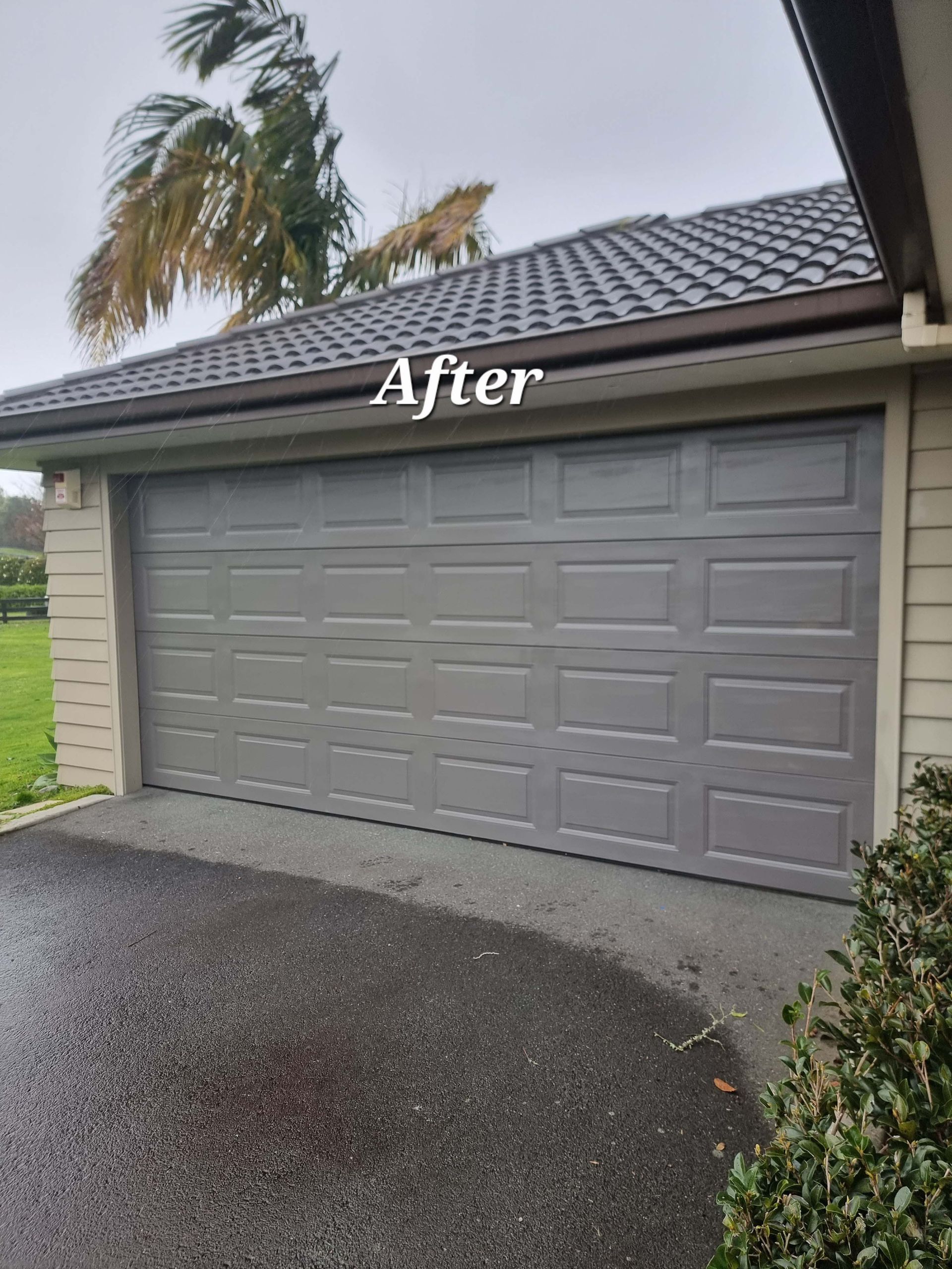 Gray double garage door on a house, labeled “After,” with a tiled roof and wet driveway.
