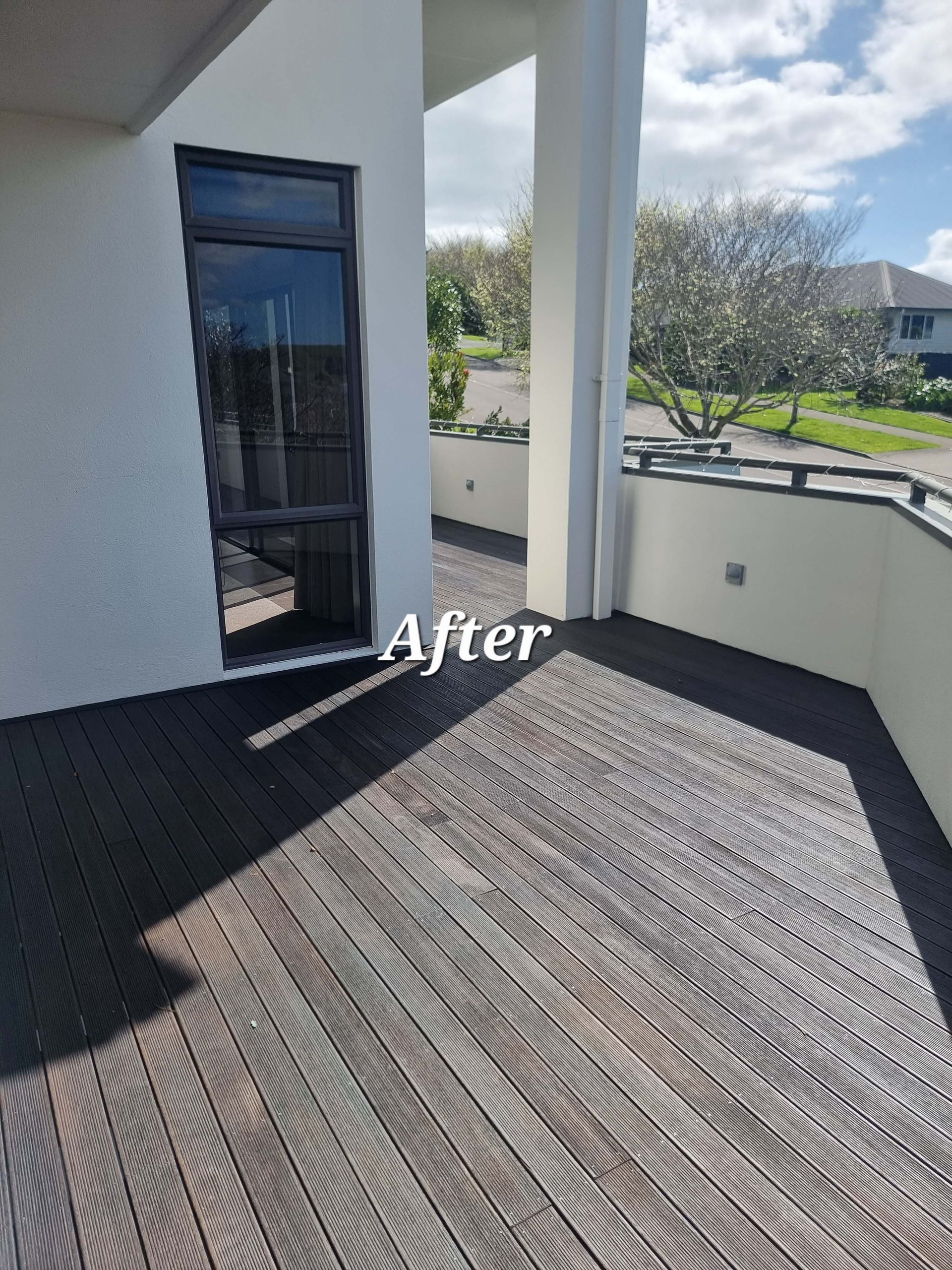 After view of a small balcony with dark decking, white walls, and a glass door in bright sunlight
