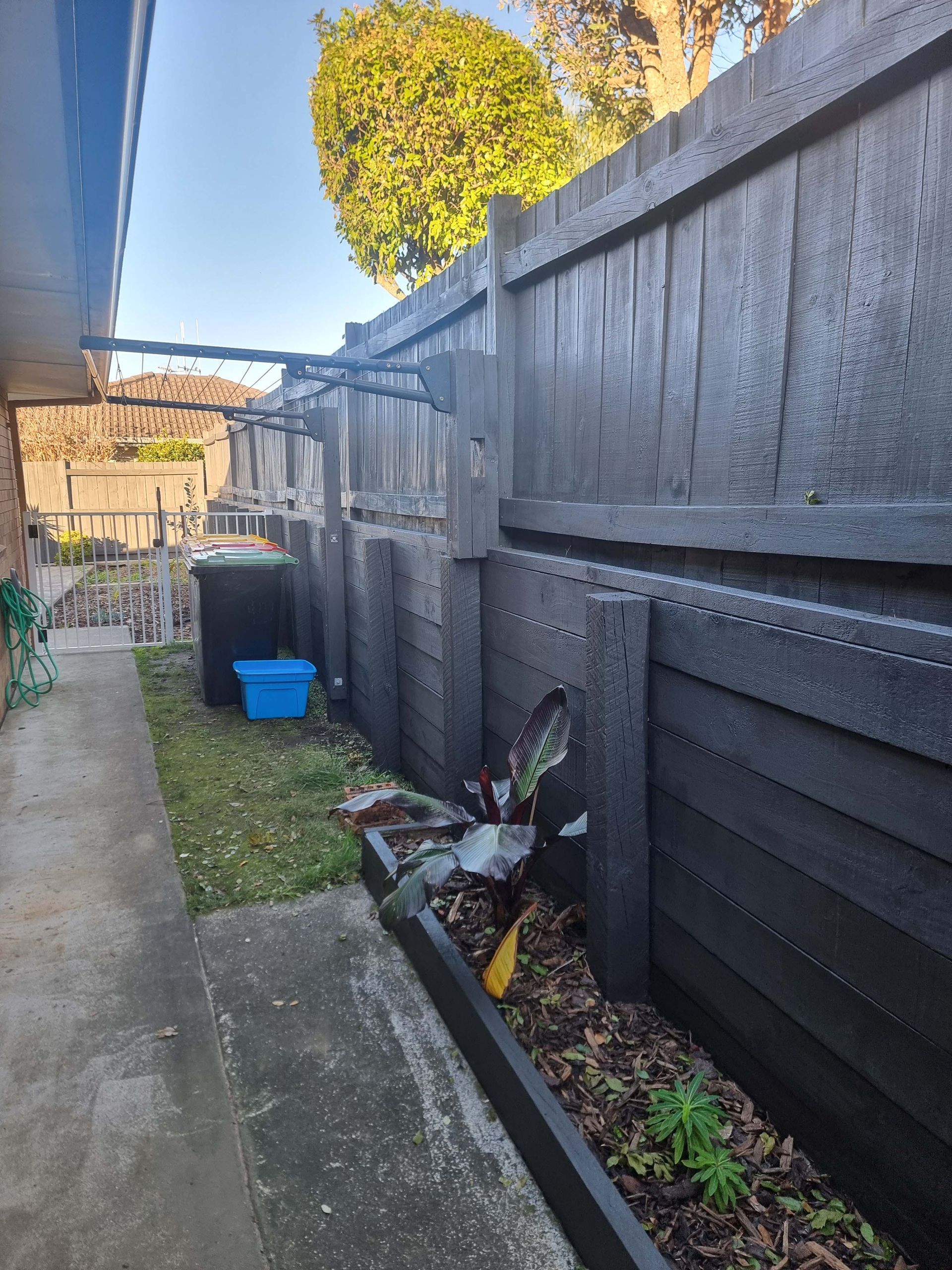 Narrow side yard with concrete path, black fence, blue bucket, and scattered debris along the wall.