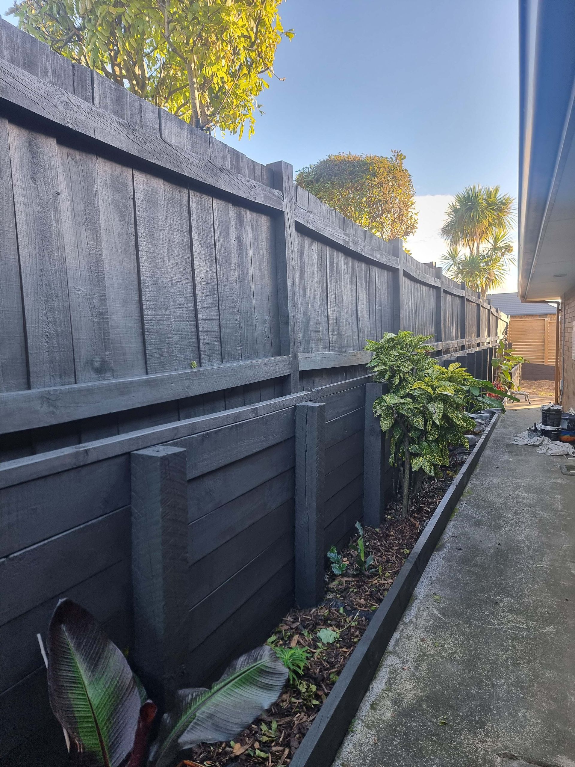 Narrow outdoor walkway beside a tall black fence, with potted plants and trees in sunlight