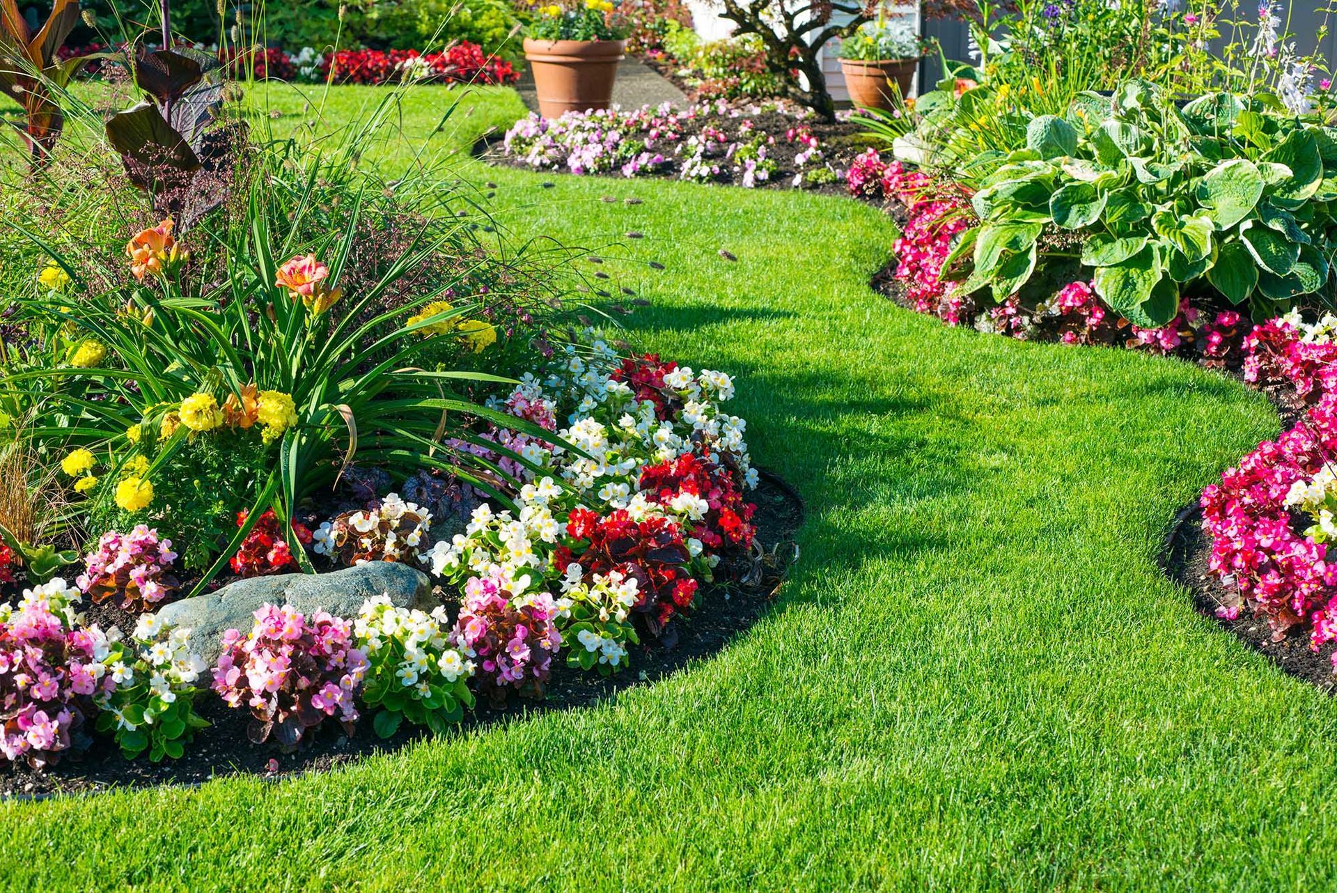 Colorful flower beds border a lush green garden path with potted plants in the background