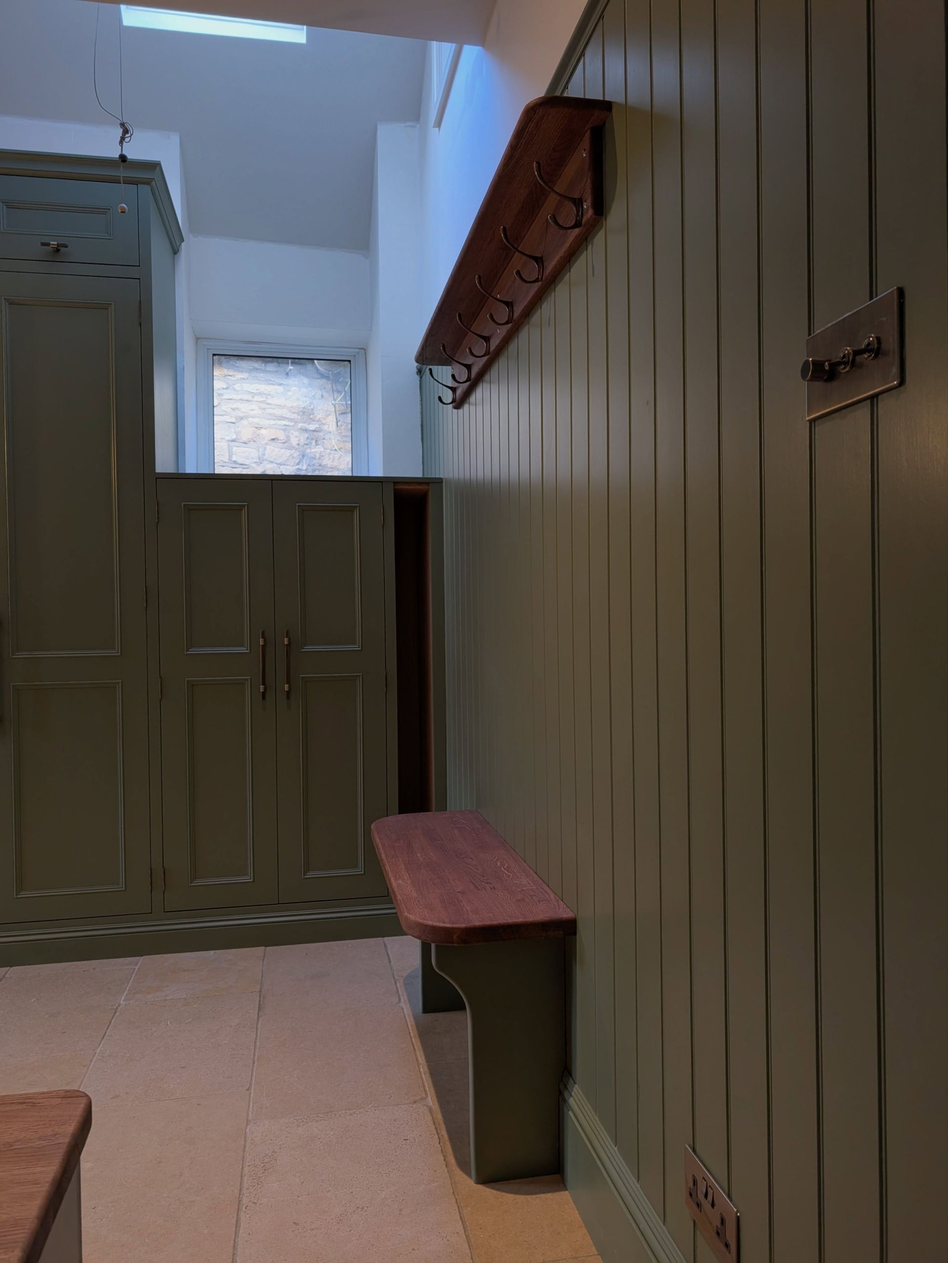 Narrow indoor hallway with green paneled walls, wooden benches, and a half-height partition leading to a bright window.