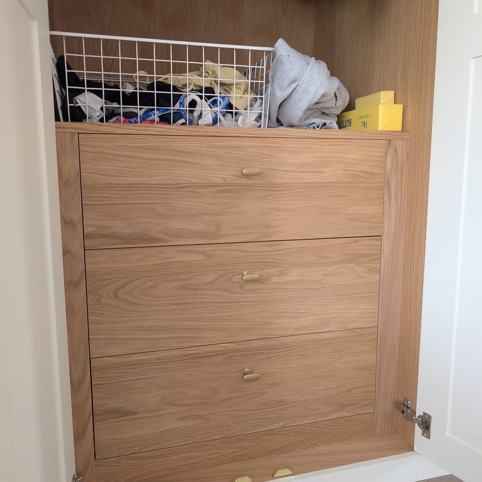 Wooden closet with two drawers and open storage bin holding folded clothes and items.