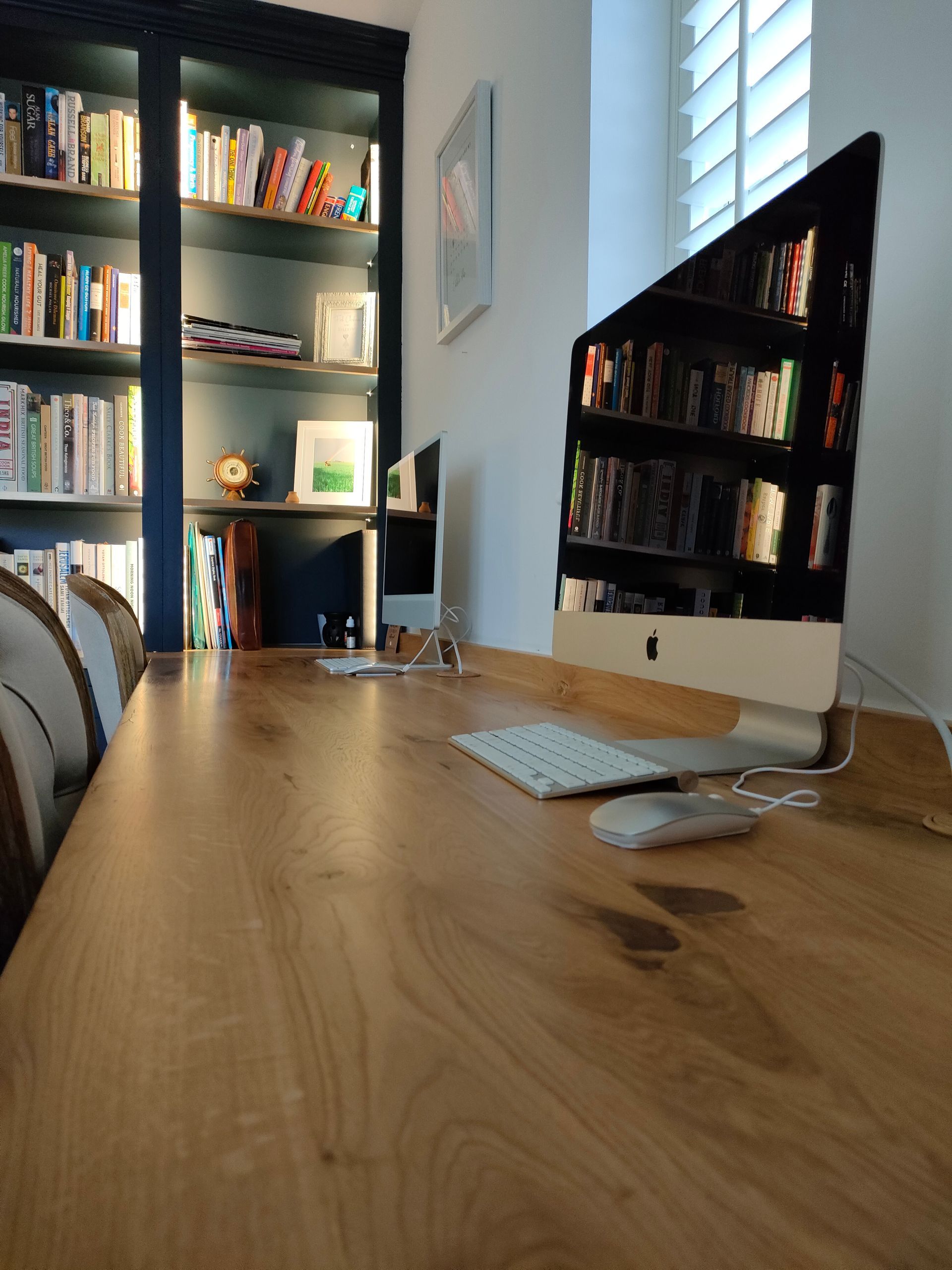 A long wooden desk with two Apple computers, white keyboards, and mice, set against a dark blue bookshelf filled with books.