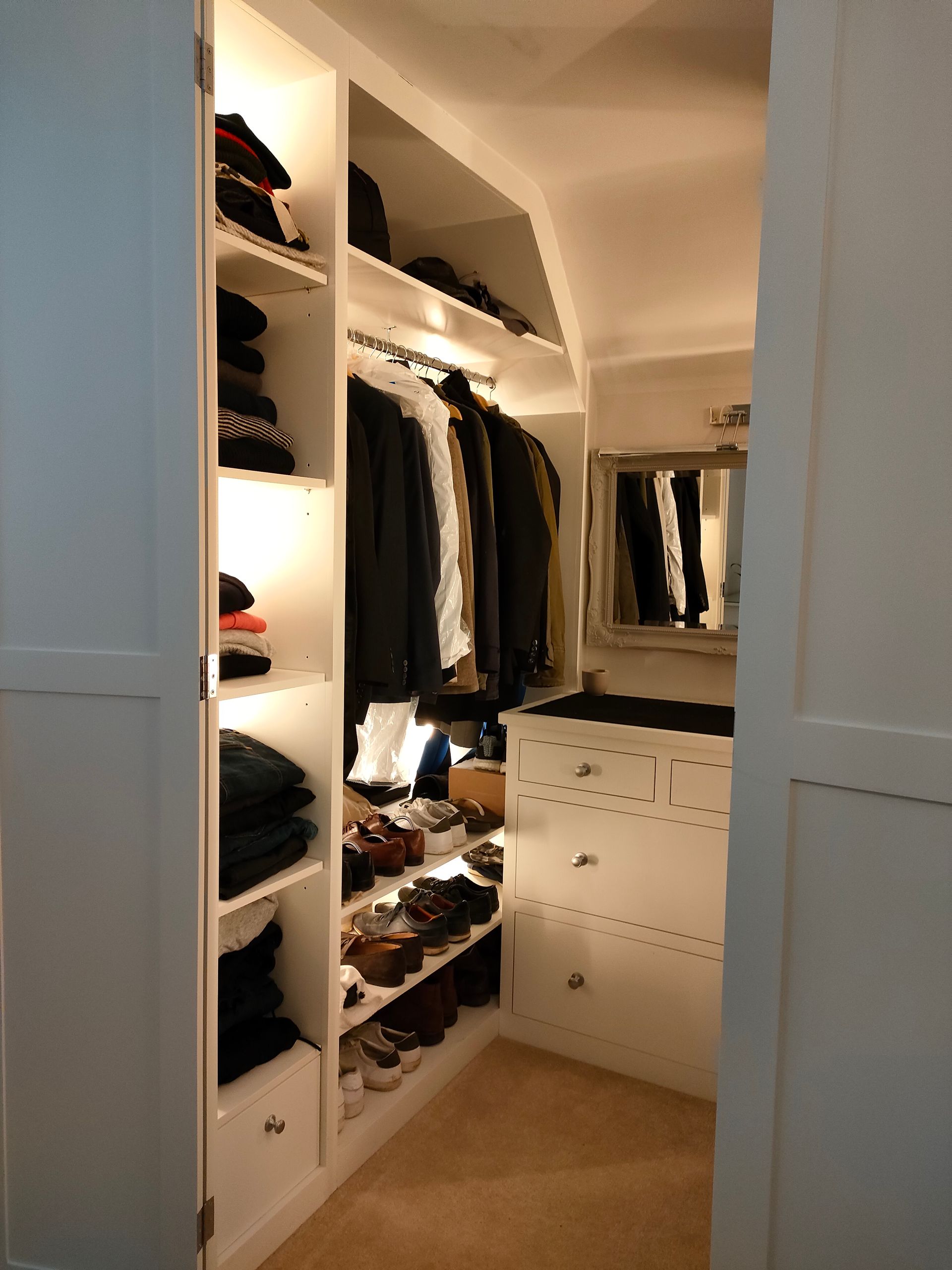 A walk-in closet with white shelving, illuminated clothing racks, stacked folded clothes, shoes, and a dresser.