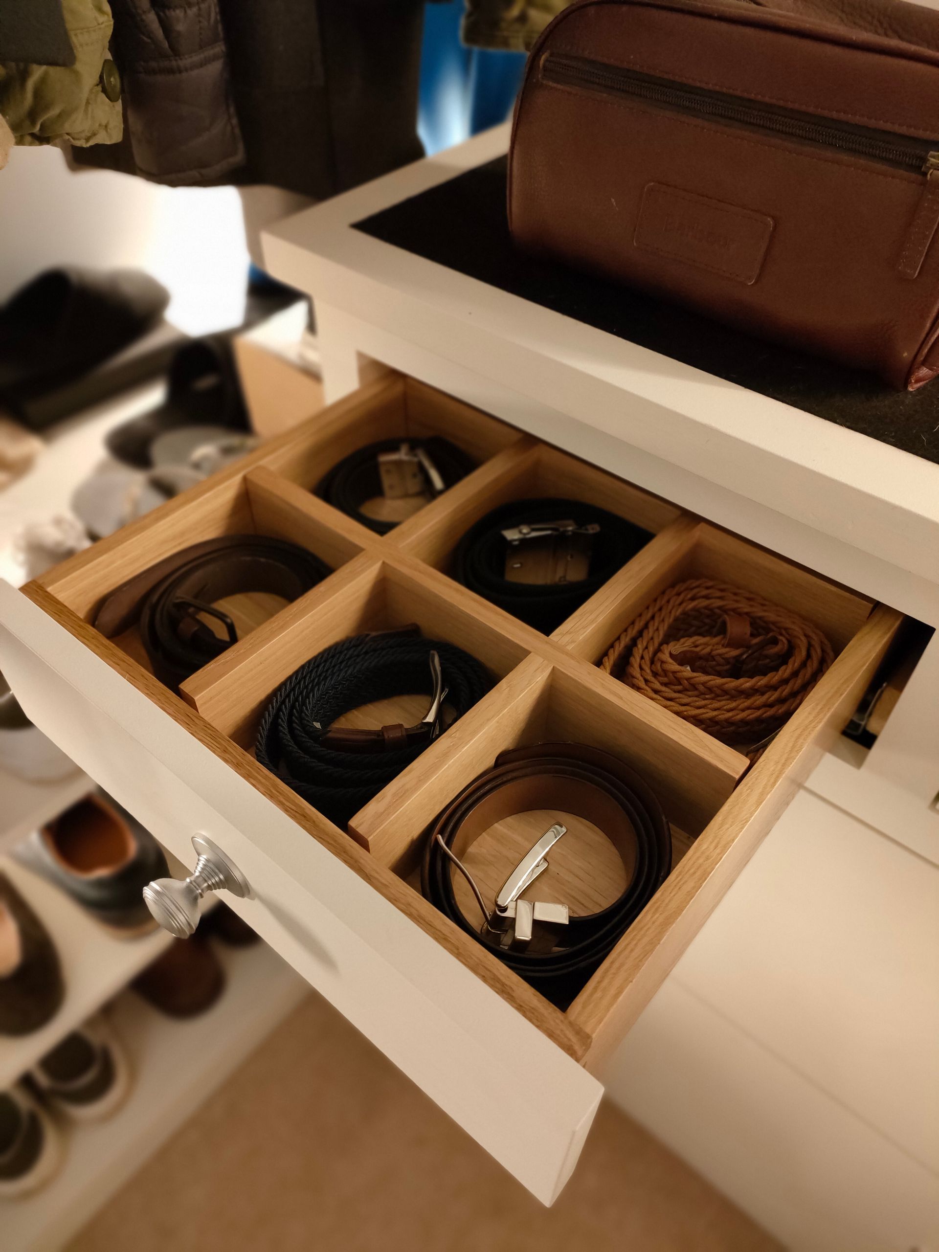 A partially open white closet drawer with six compartments, each holding a neatly coiled leather belt.