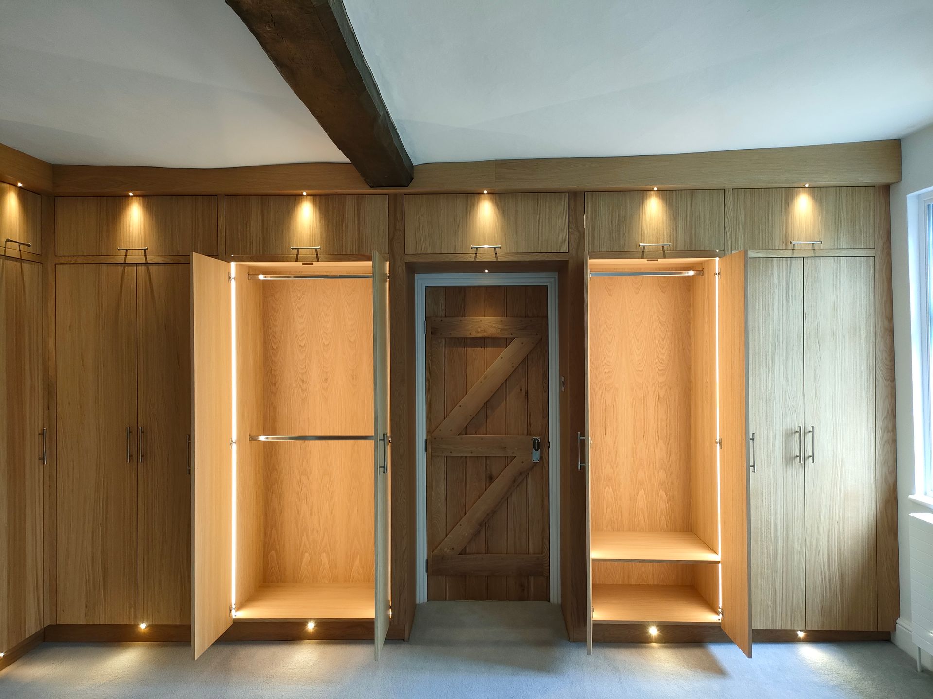 Built-in wooden wardrobes surround a central wooden door, featuring illuminated interiors and recessed overhead lighting.
