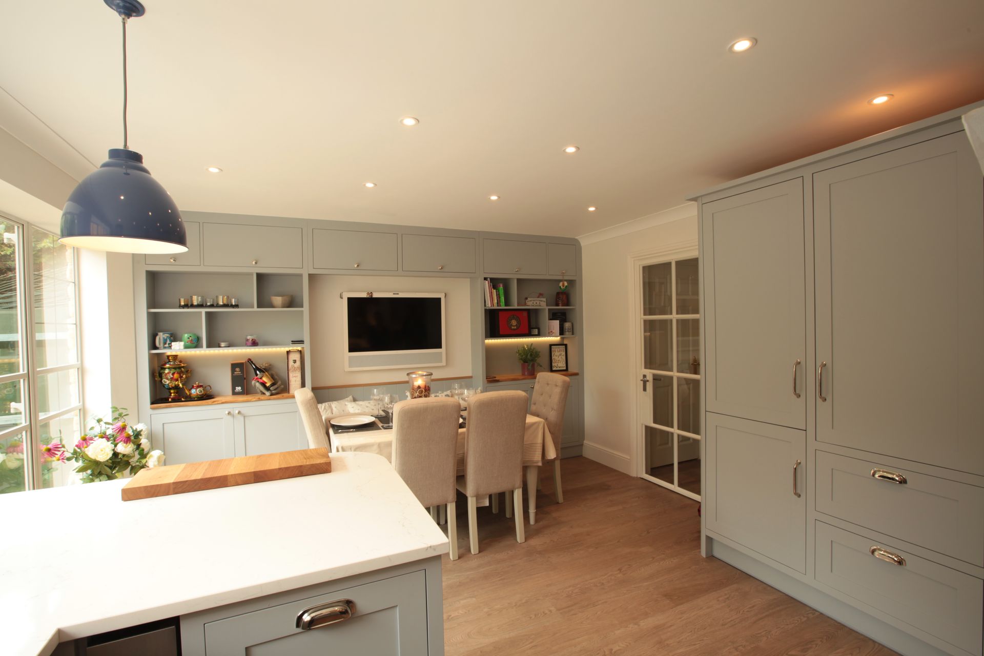 A bright, modern kitchen and dining area with sage-green cabinets, a center island, and a dining set near a wall-mounted TV.