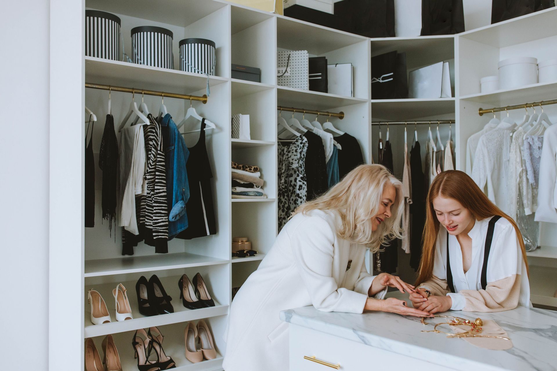 Two women browsing accessories in a bright walk-in closet with shelves of clothes and shoes