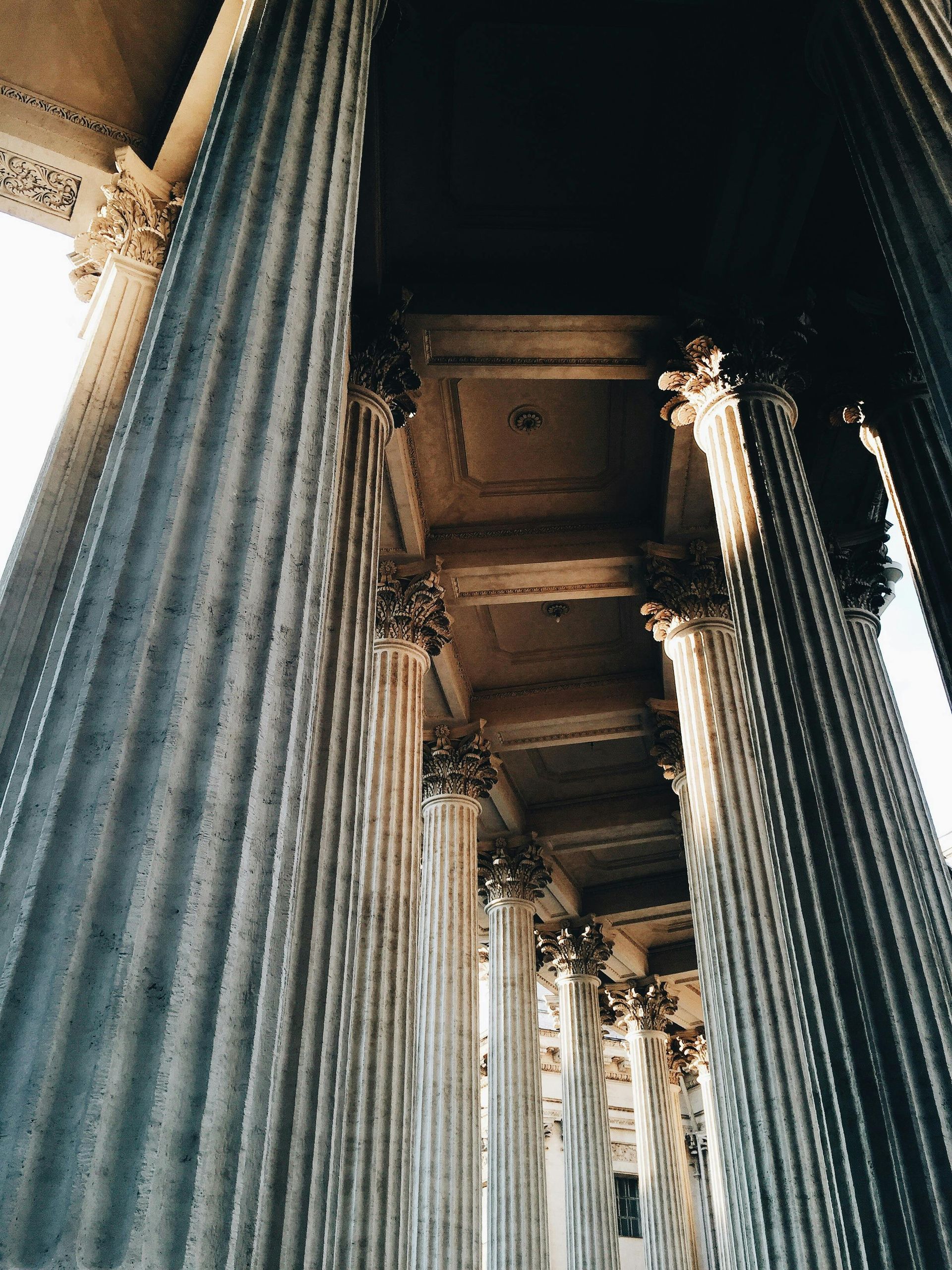 A low-angle view looking down a colonnade of tall, fluted stone pillars with ornate capitals against a shadowed ceiling.