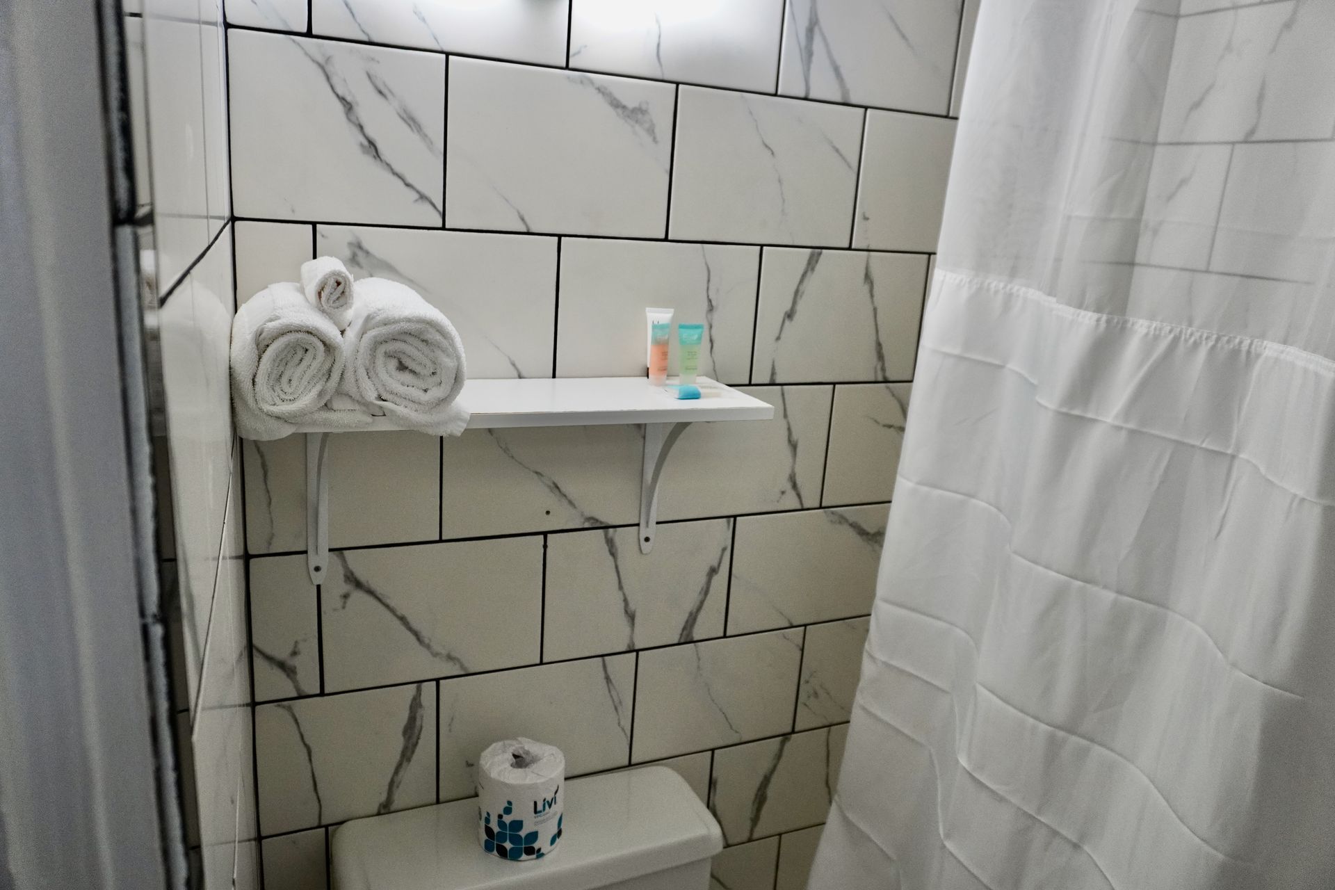 A bathroom with a toilet , shower curtain , shelf and towels.