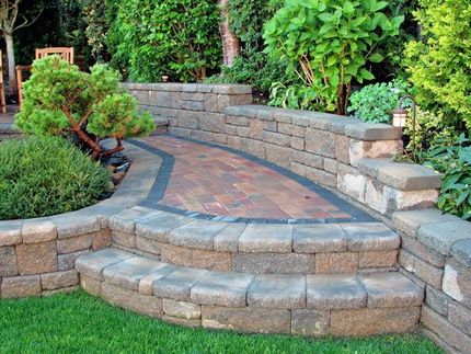 Curved stone steps and a brick patio bordered by retaining walls and lush green landscaping