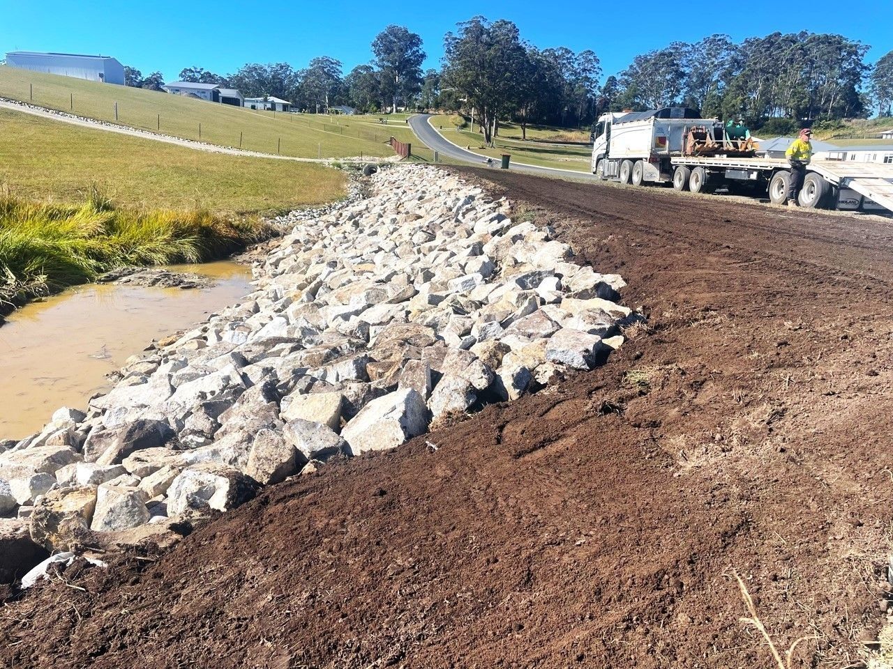 Rocks Lining a Creek Bed Next to a Dirt Road, With a Truck and Worker — Blue Dog Earthmoving Landscape Supplies in Newee Creek, NSW