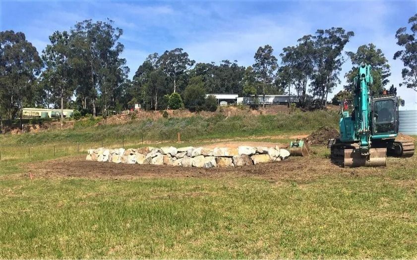 An Excavator Near a Rock Structure on a Grassy Hillside — Blue Dog Earthmoving Landscape Supplies in Newee Creek, NSW