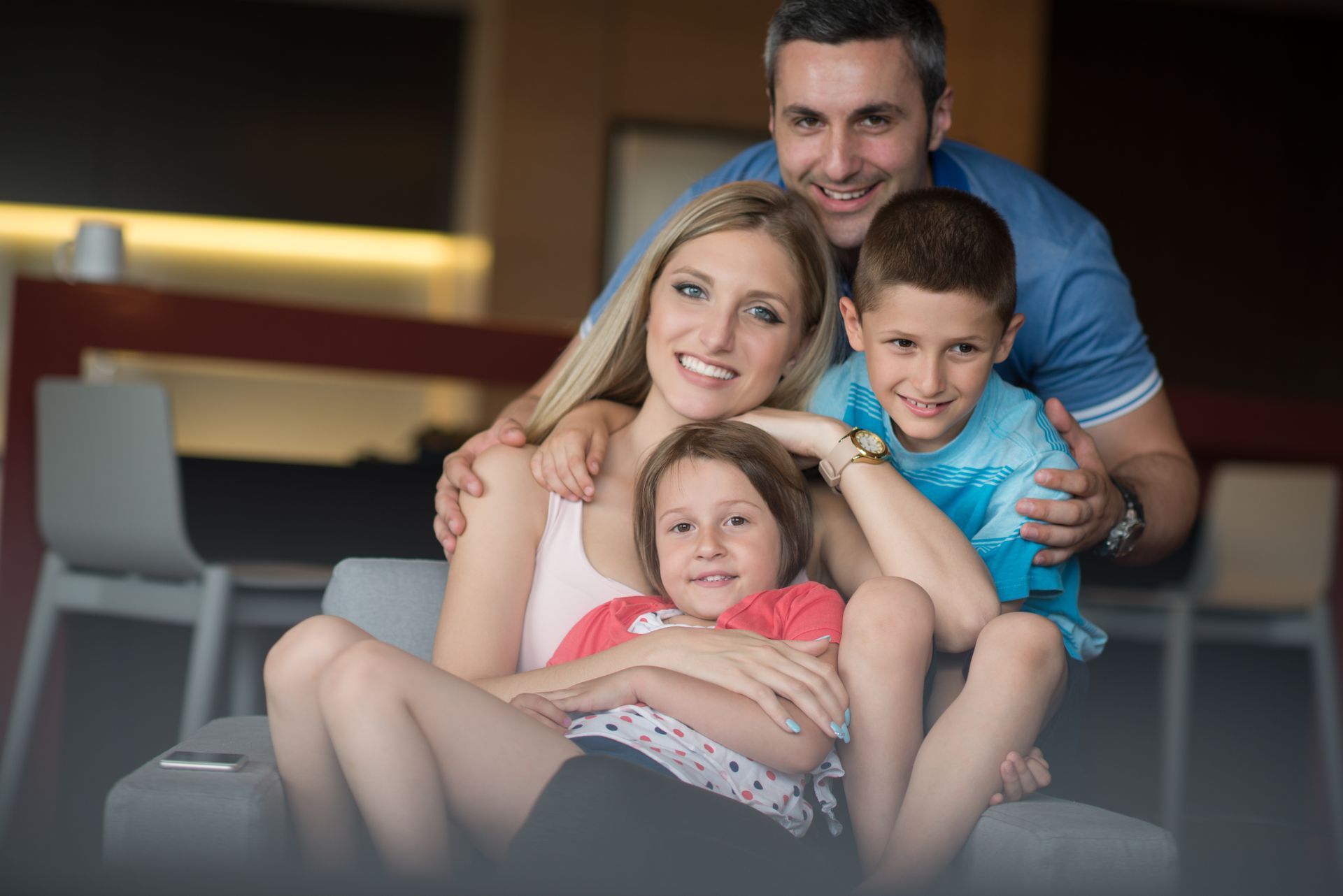 Family of four smiling together on a couch indoors, with parents embracing two children.