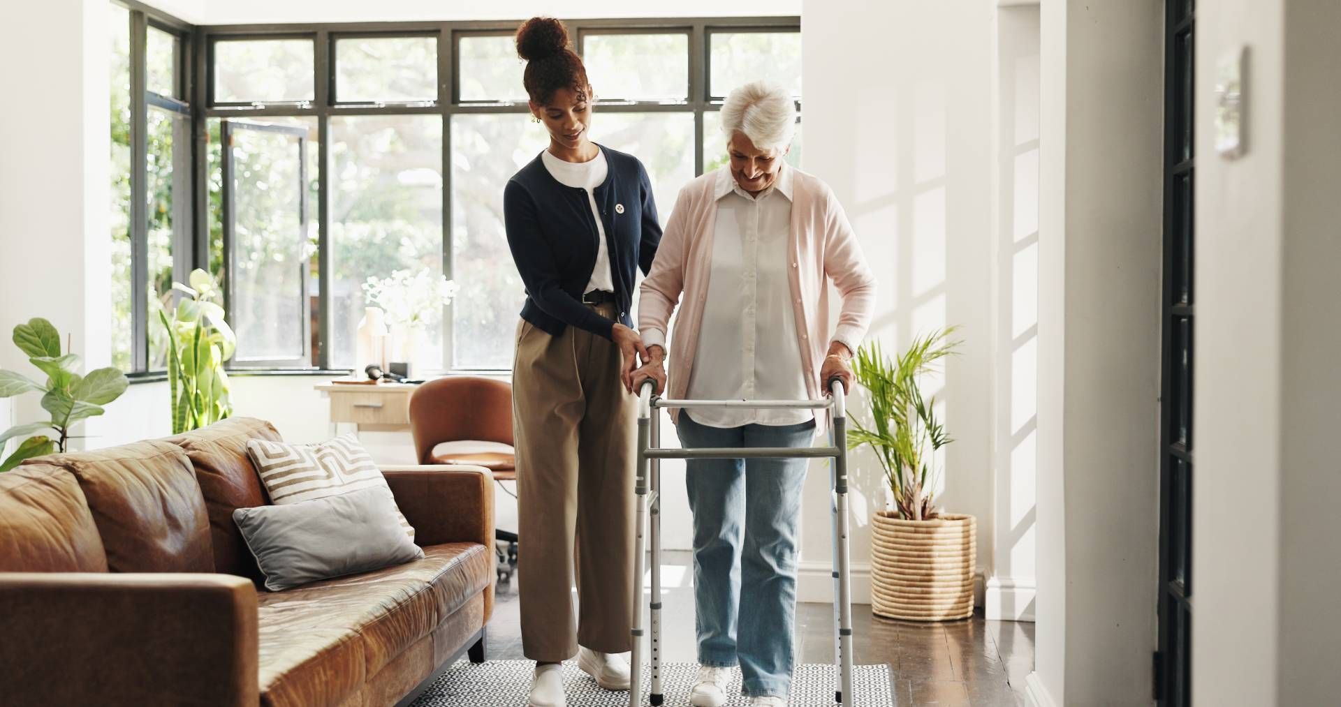 Two people using a walker in a bright living room with a sofa and large windows