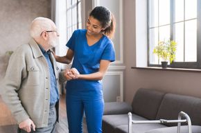 Caregiver in blue scrubs helping an older man stand in a bright living room