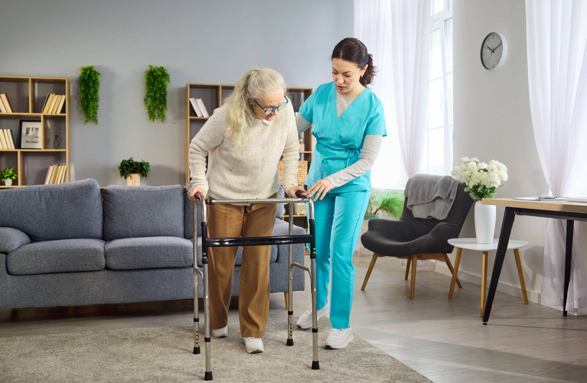 Caregiver assists an elderly person walking with a walker in a bright living room