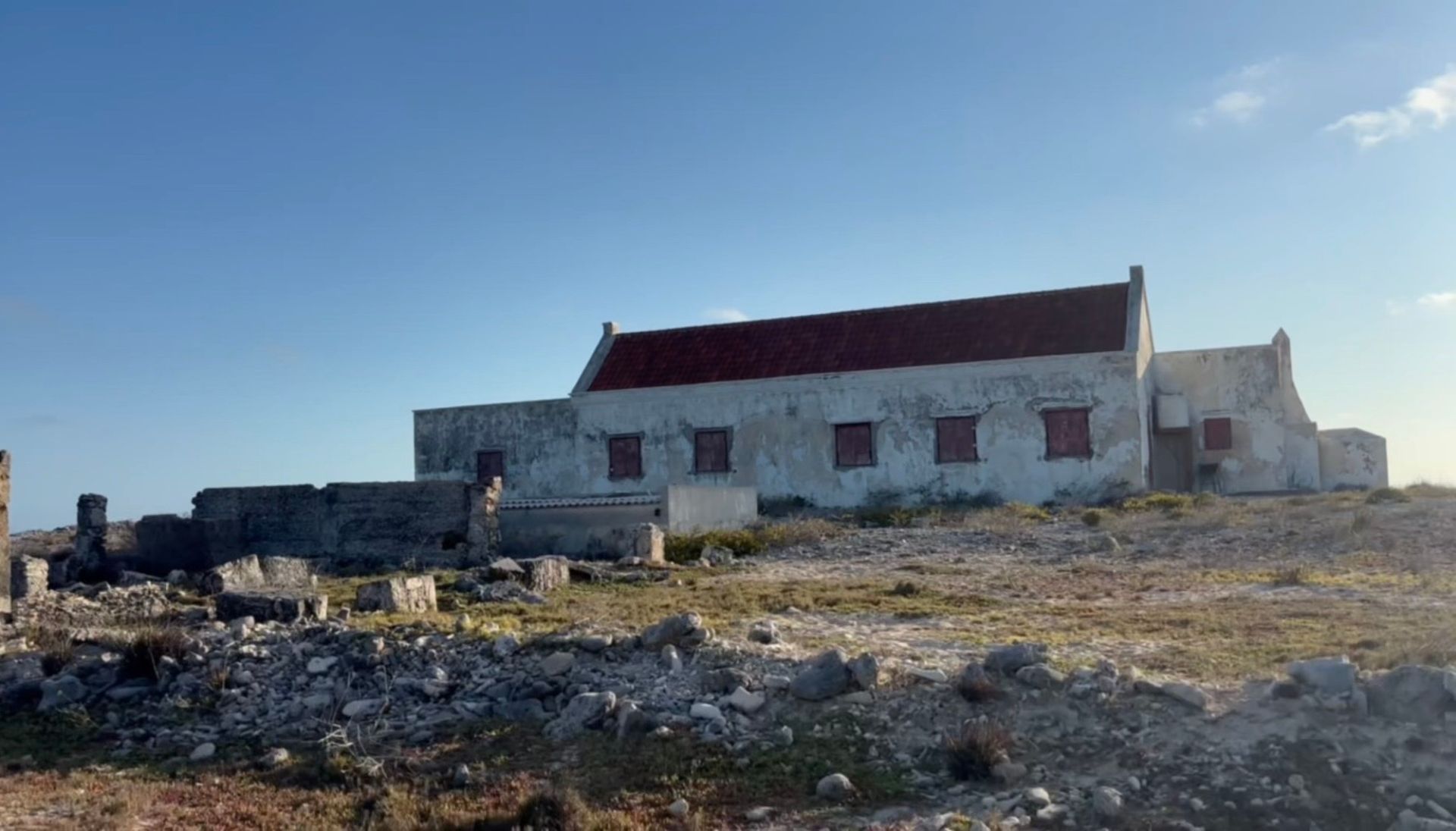 Small white stone building Bonaire with a red roof on a rocky hillside under a clear blue sky