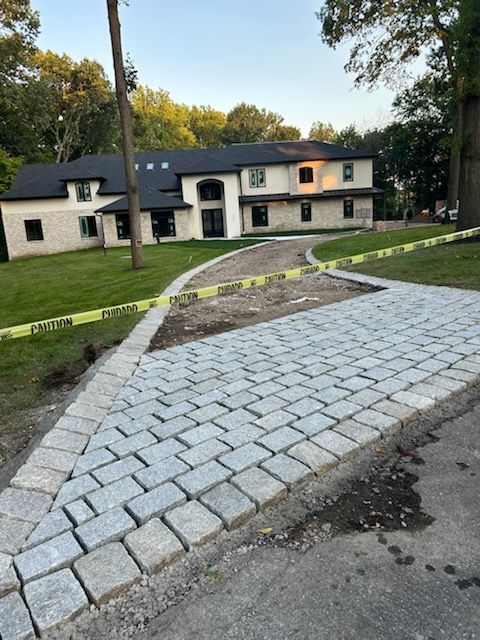 Completed cobblestone driveway leads to a two-story beige house with black-framed windows. Yellow caution tape.