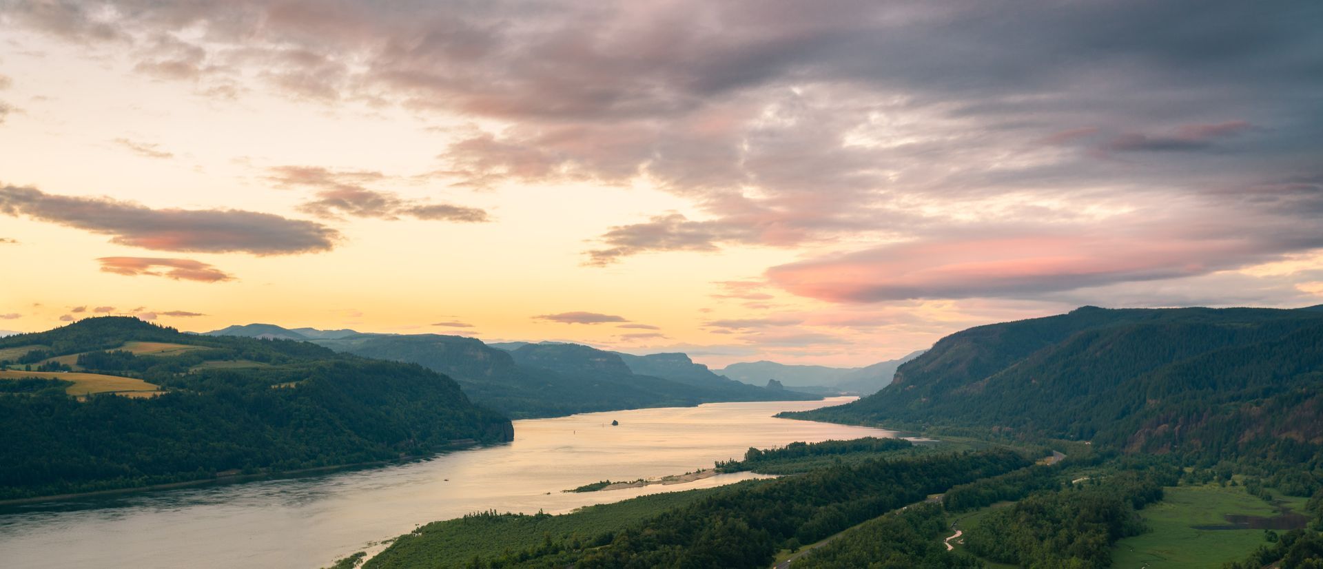 A river winds through a lush valley, with forested hills and a colorful sunset sky overhead.