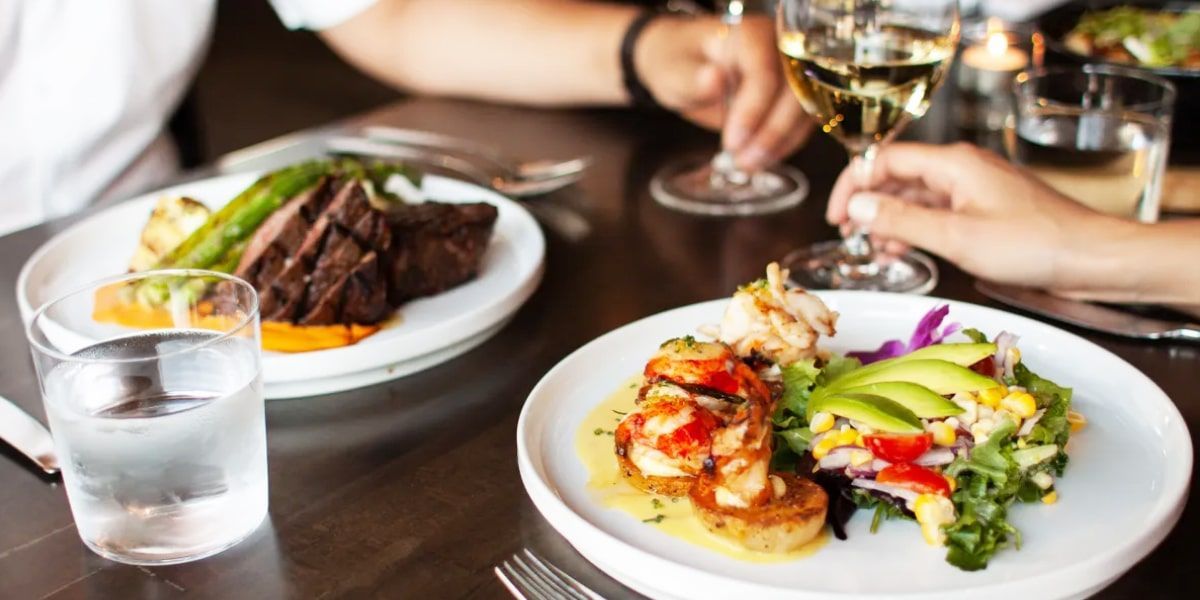 Two plates of food and two glasses of wine on a table. One plate has steak and asparagus. Another has shrimp and salad.