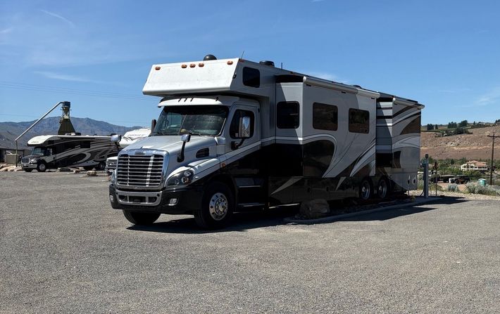 White RV parked in a bright desert landscape; person near back opening compartment, skis leaning nearby.
