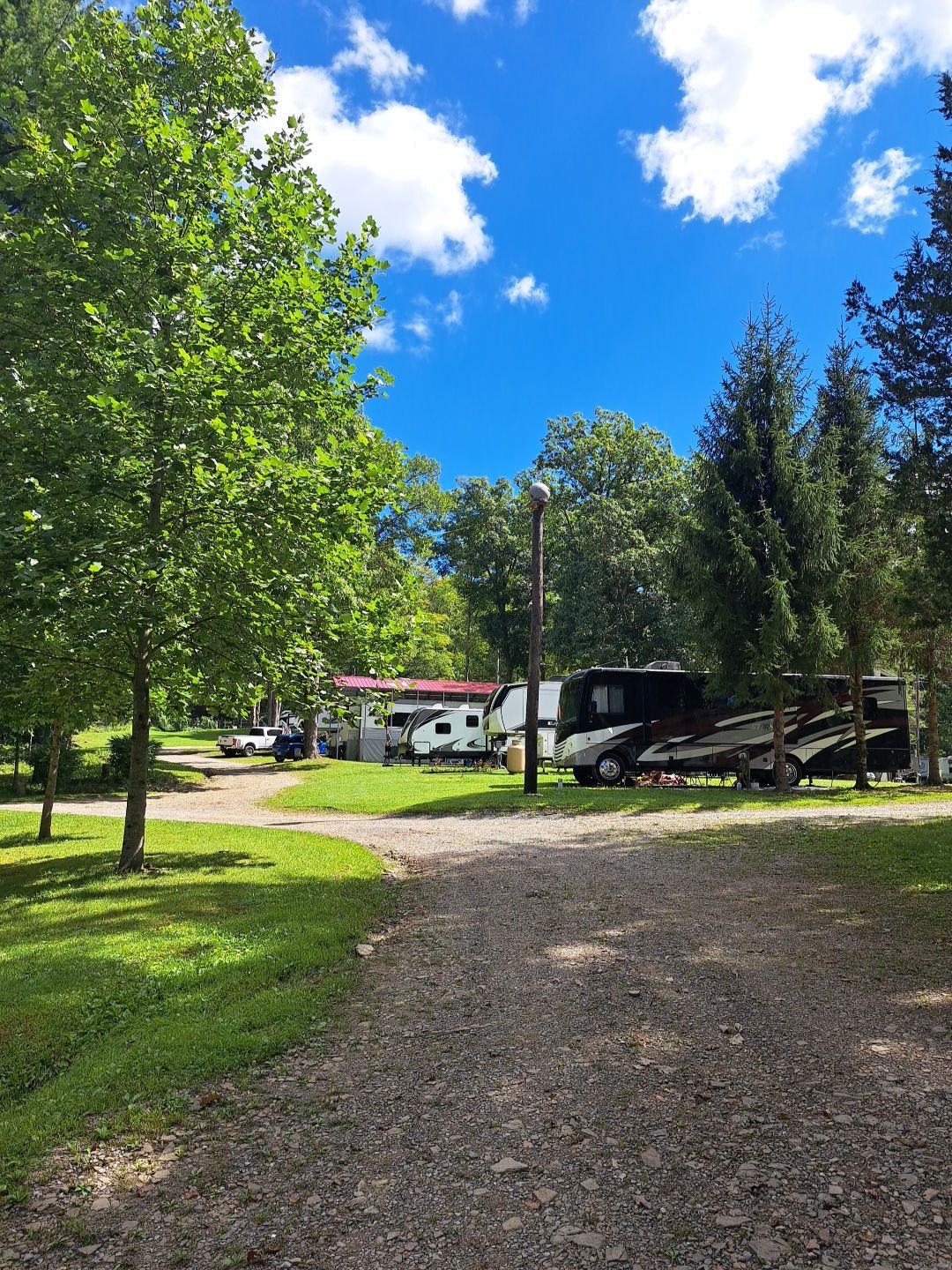 Gravel driveway leading to a white house and RVs, framed by green trees under a bright blue sky.