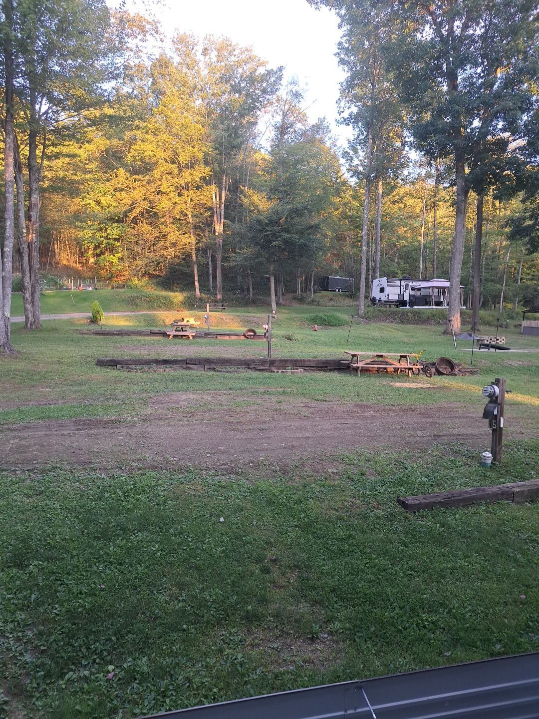Wooded campsite with picnic tables and a trailer, lit by warm evening light.