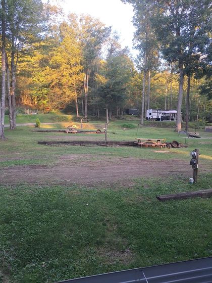 Wooded campsite with picnic tables and a trailer, lit by warm evening light.