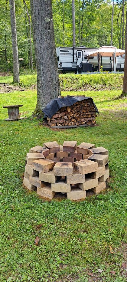Circular brick fire pit with stacked firewood in a grassy yard, near a tree and a covered wood pile.