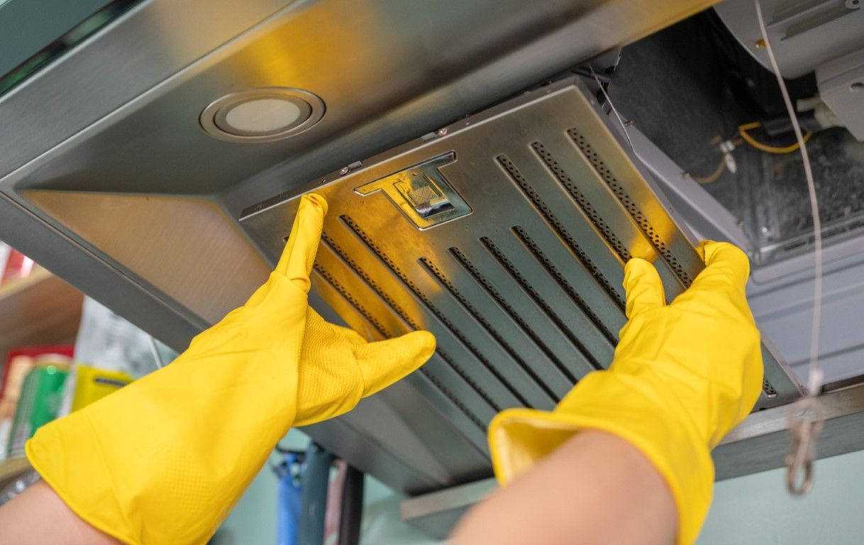 Yellow-gloved hands removing a metal range hood filter from a kitchen exhaust hood