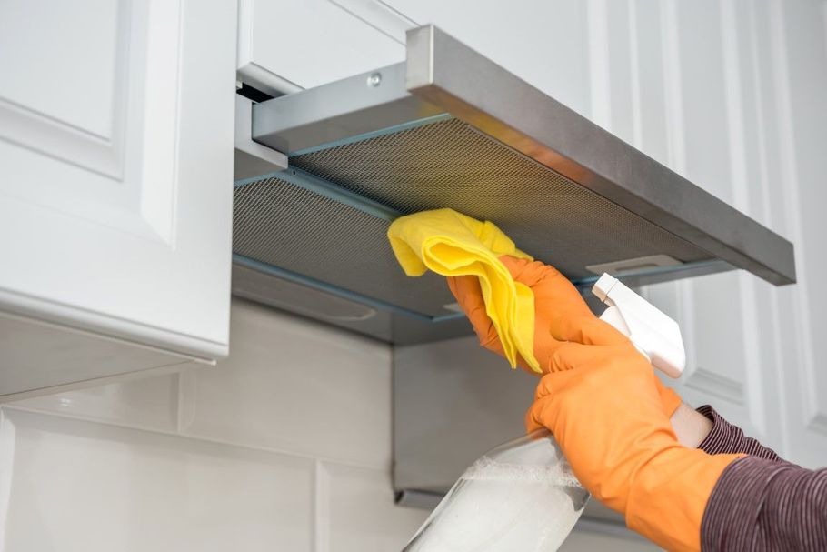 Person cleaning a kitchen range hood with yellow cloth and spray bottle, wearing orange gloves