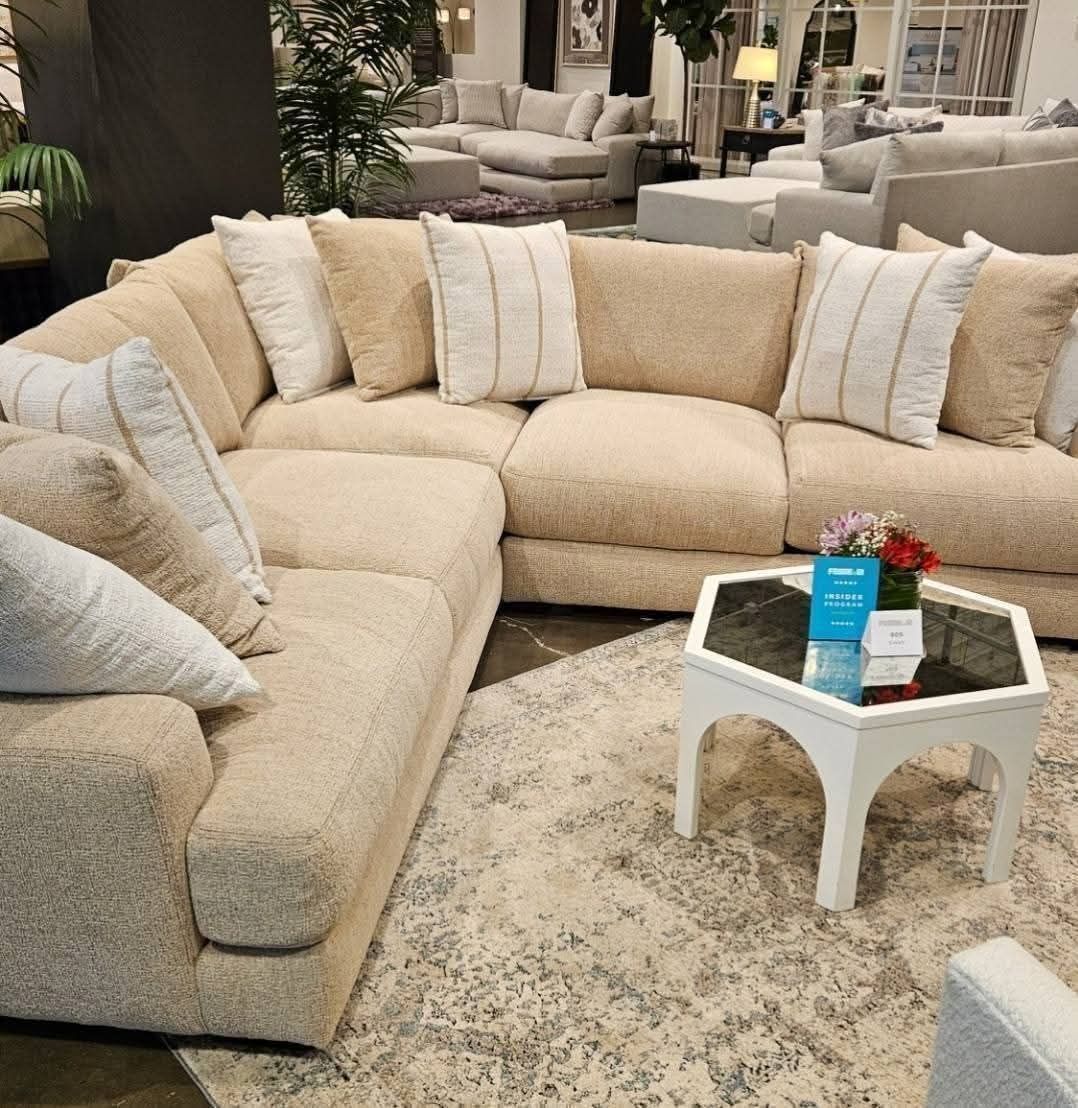 Beige sectional sofa with pillows in a furniture store setting. White hexagon coffee table and patterned rug.