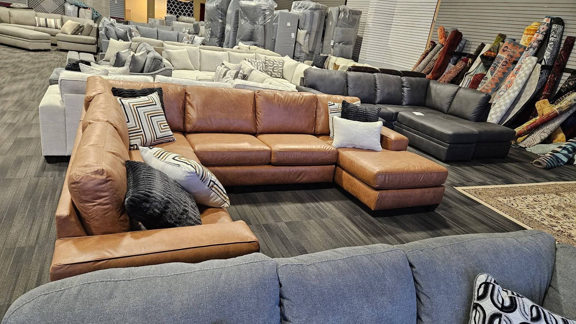 Brown leather sectional sofa displayed in a furniture store, surrounded by other couches and rugs.