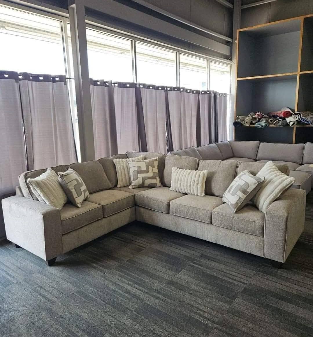 Beige sectional sofa with patterned throw pillows in a room with gray curtains and shelves.