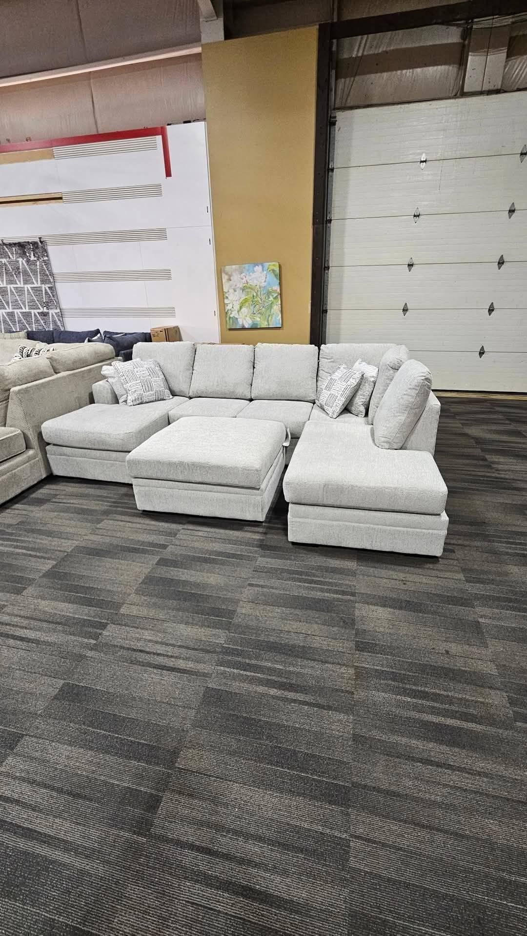 Light gray sectional sofa with ottoman in warehouse setting.