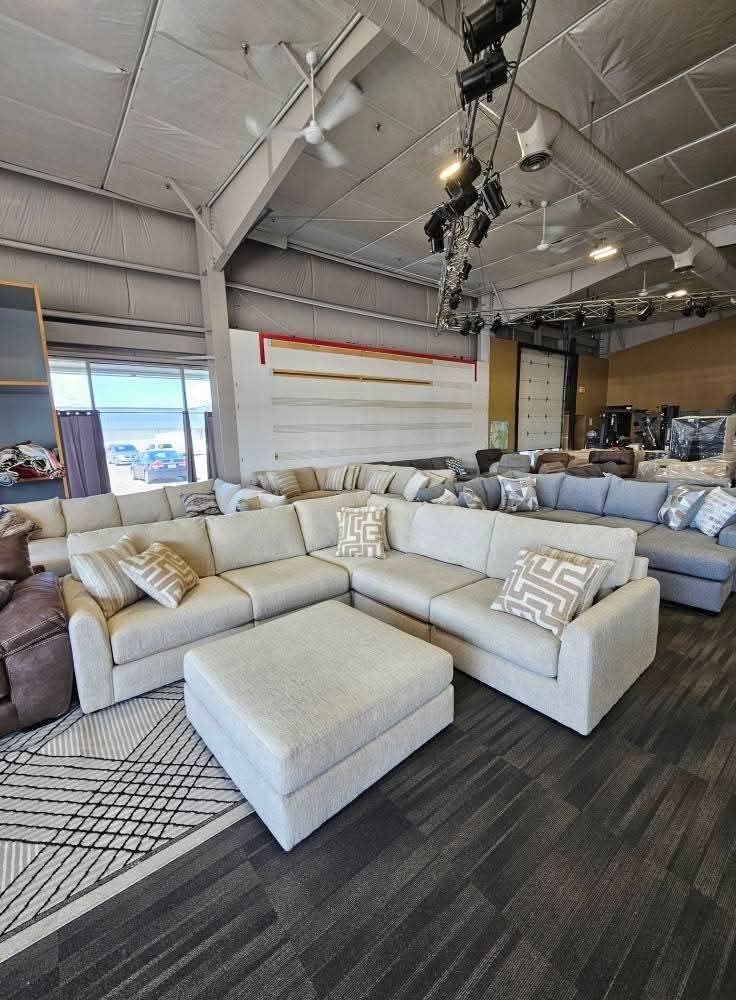 A furniture store showroom with a light gray sectional sofa and matching ottoman. Dark carpet.