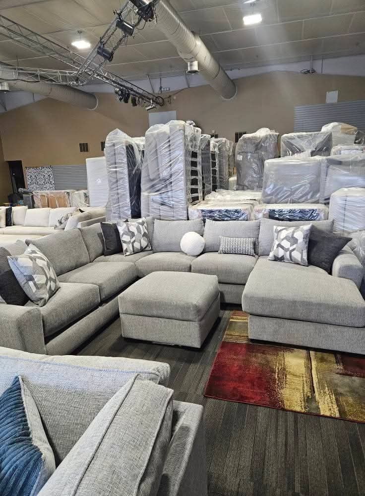 Warehouse interior with a gray sectional sofa, ottoman, and wrapped furniture stacks.