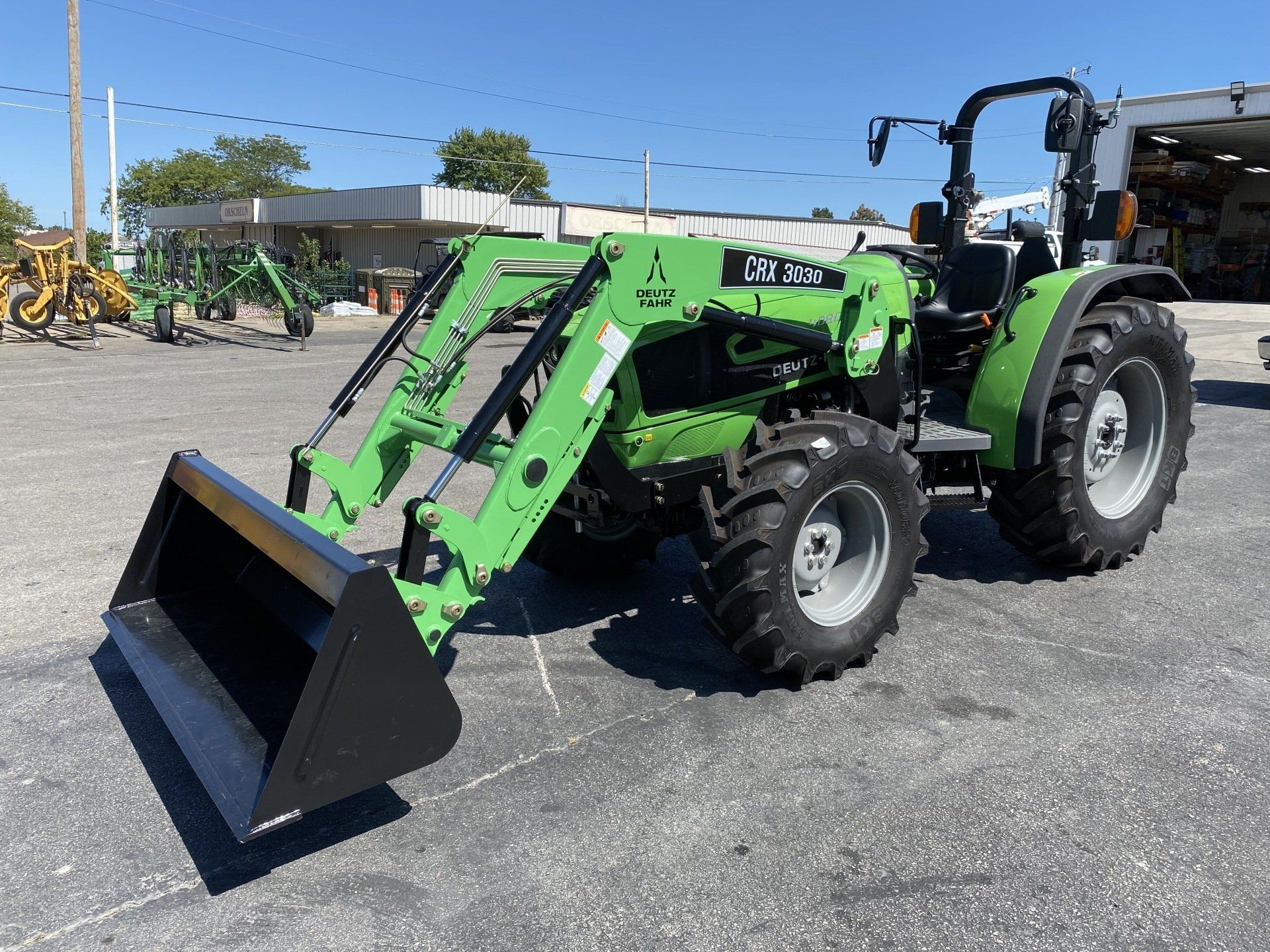 Green tractor with loader bucket on asphalt. Outdoors in front of other equipment.