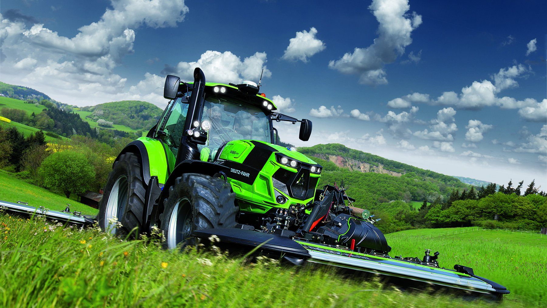 Green tractor mowing a grassy hillside under a blue sky with clouds.