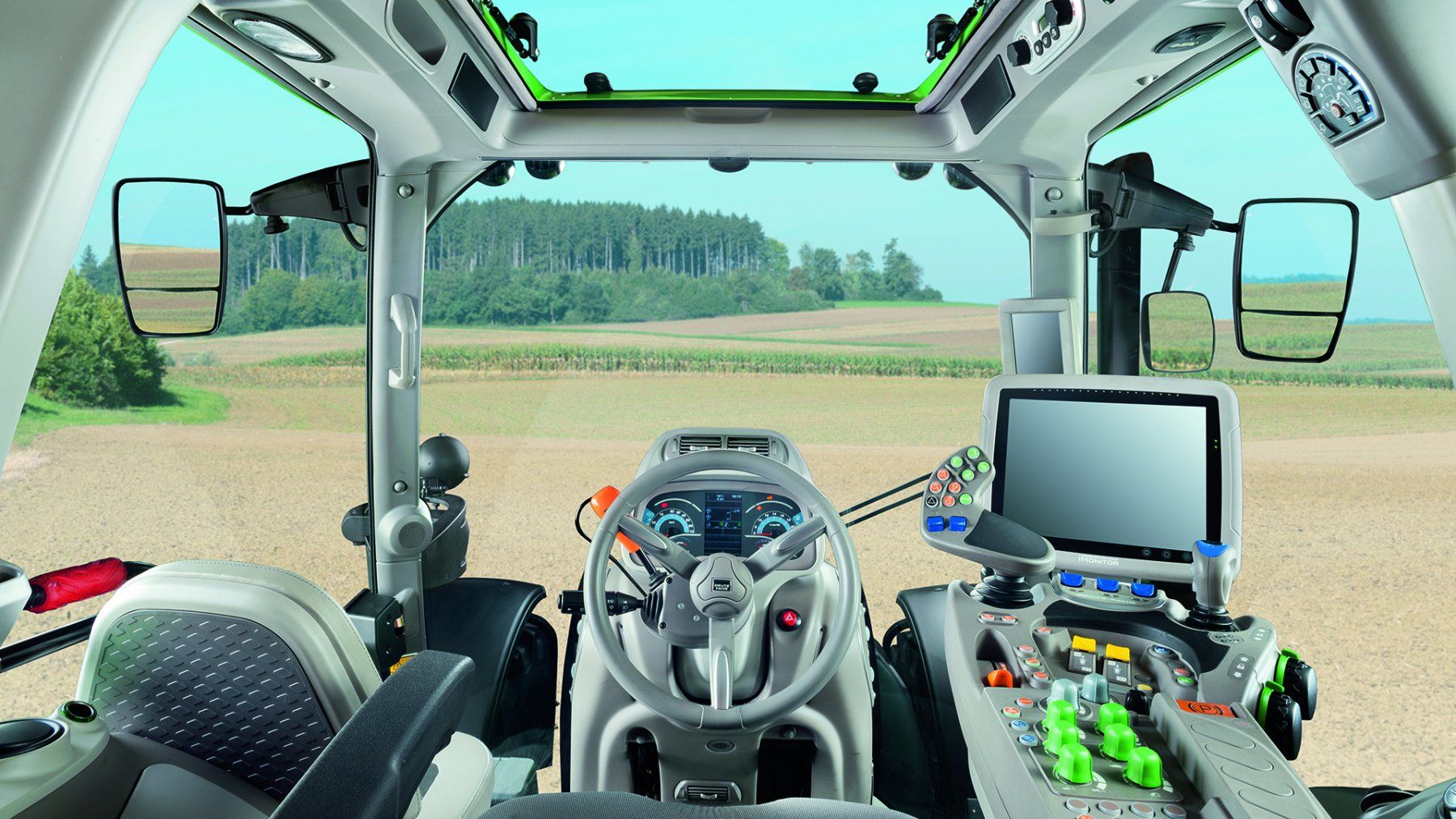 Interior view of a tractor cab with a dashboard, steering wheel, and display screen, overlooking a field.