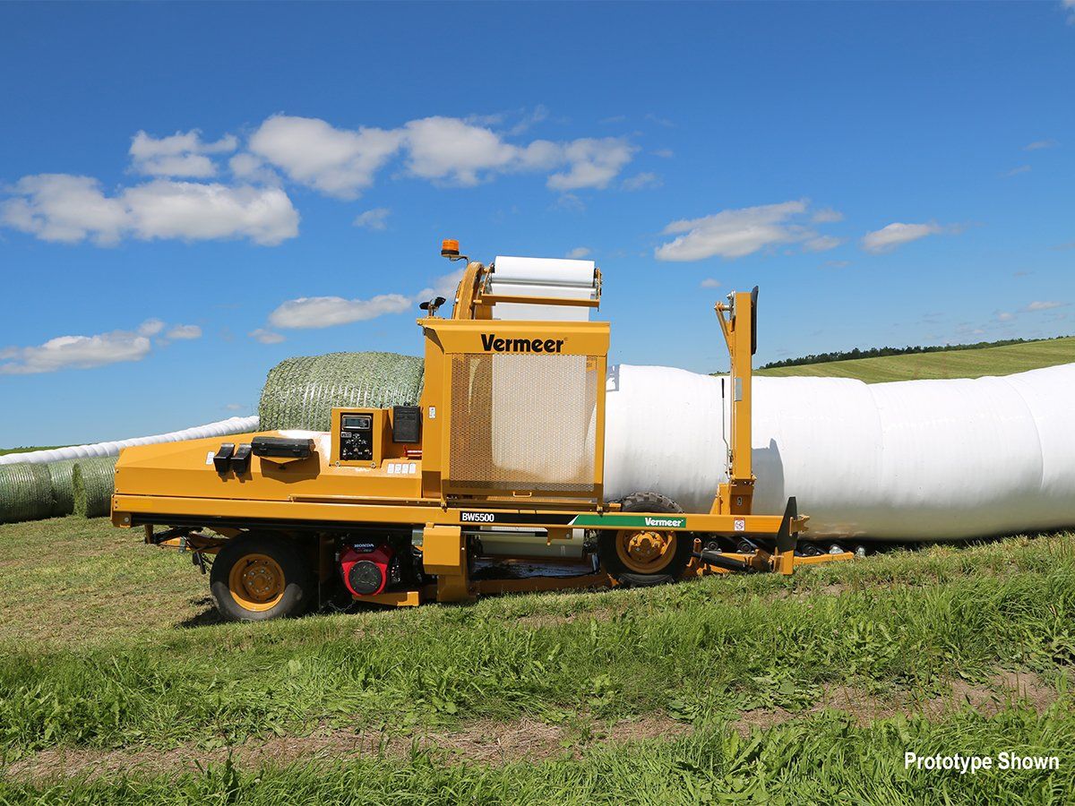 Yellow Vermeer silage bagger in a grassy field, creating a long white tube of stored feed under a blue sky.
