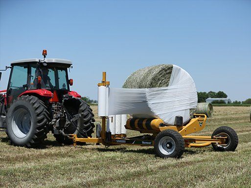 Red tractor pulling a yellow bale wrapper in a field, wrapping a round bale in white plastic.