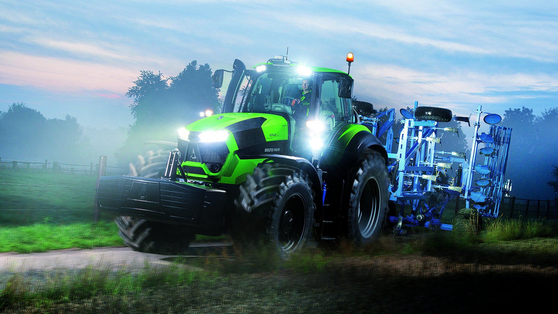 Green tractor with bright headlights and blue farm implement in a field at dusk.