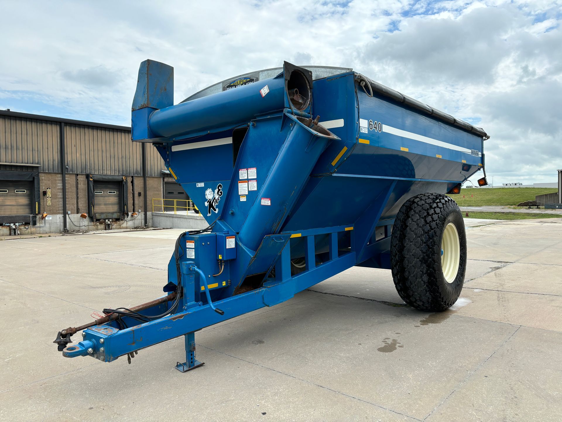 Blue agricultural grain cart on a paved surface with a building and sky in the background, front angled shot.