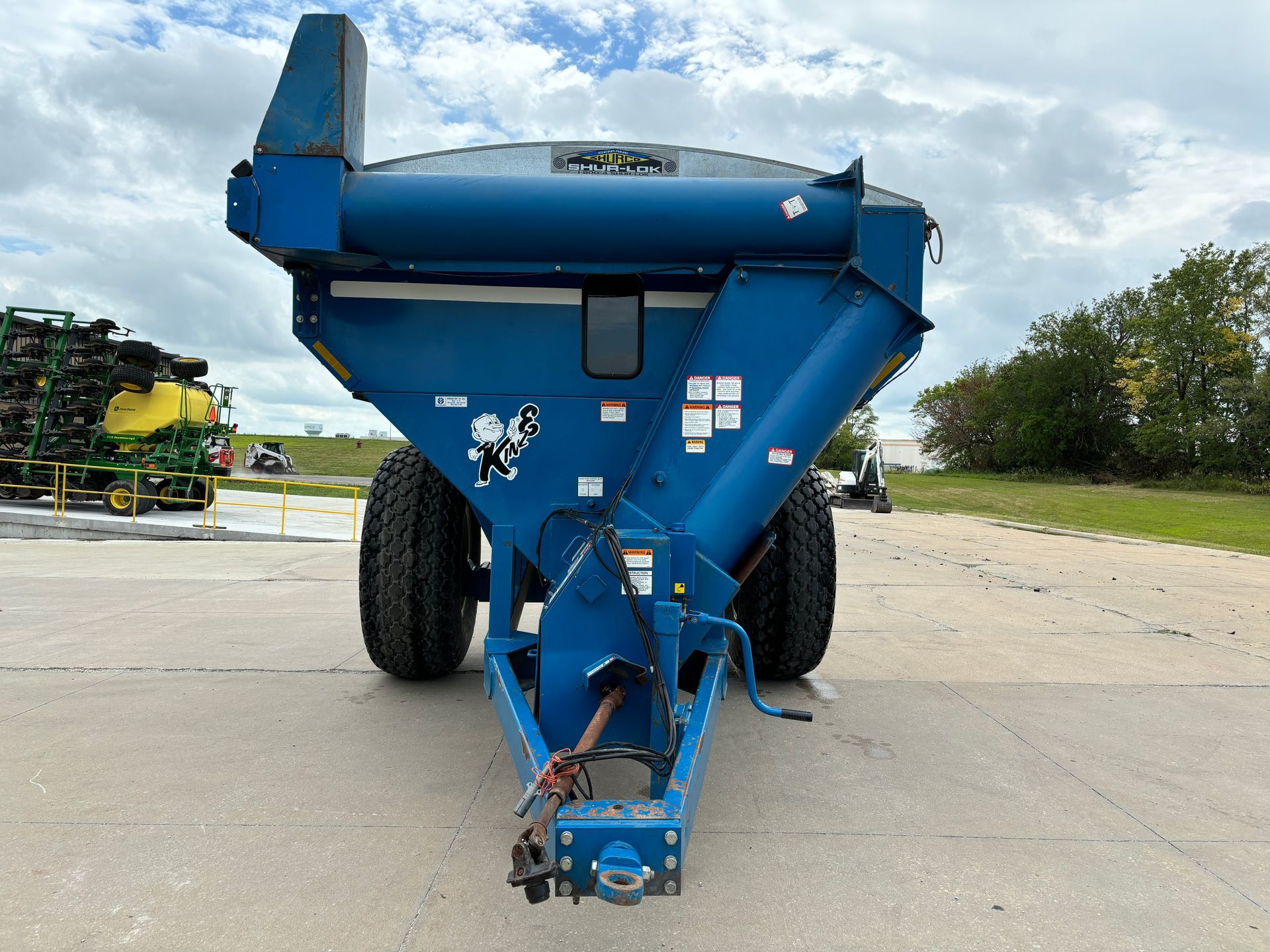 Blue agricultural grain cart on a paved surface with a building and sky in the background, front shot.