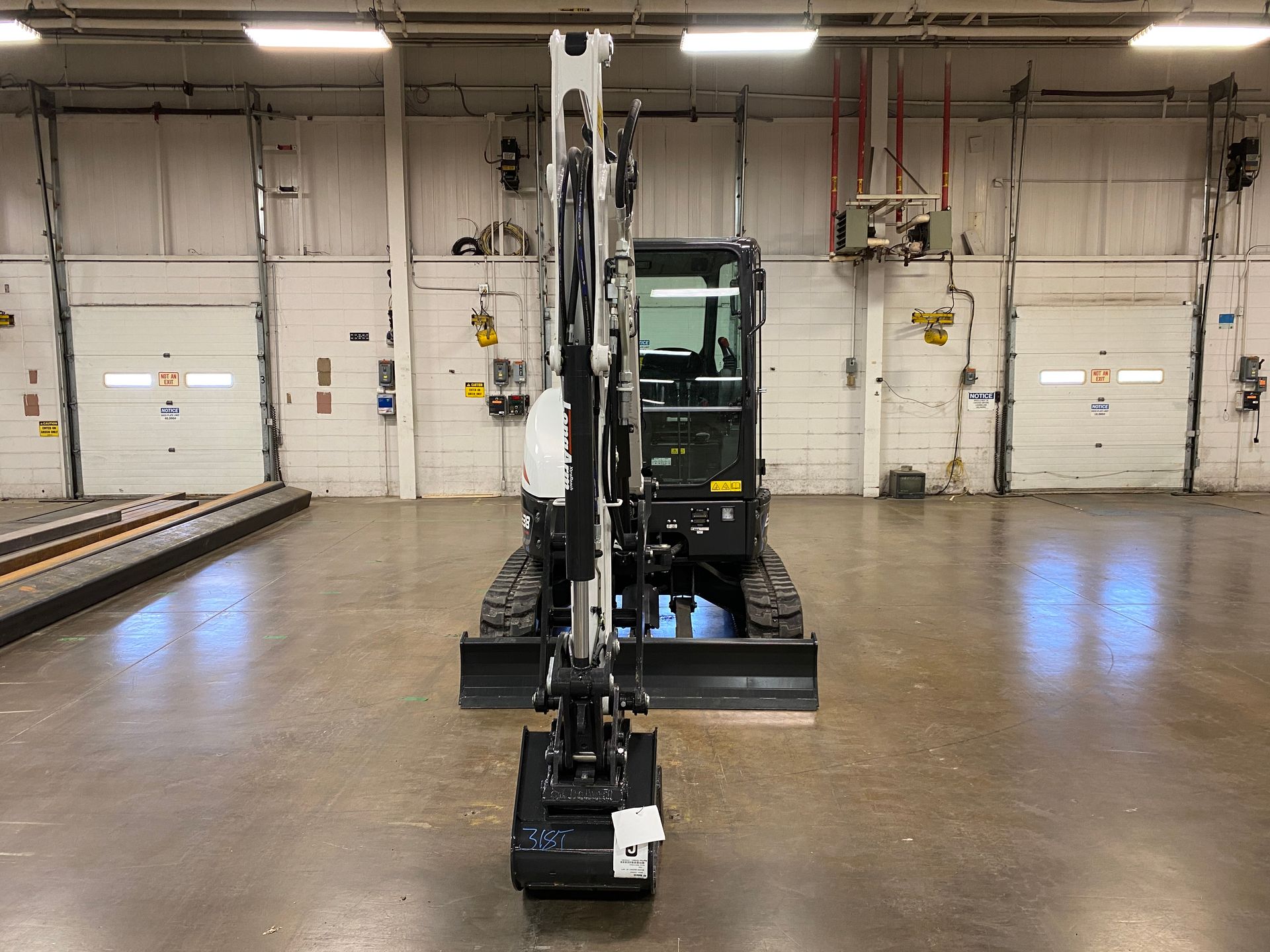 A small, white and black excavator sits in a warehouse with closed garage doors behind it.